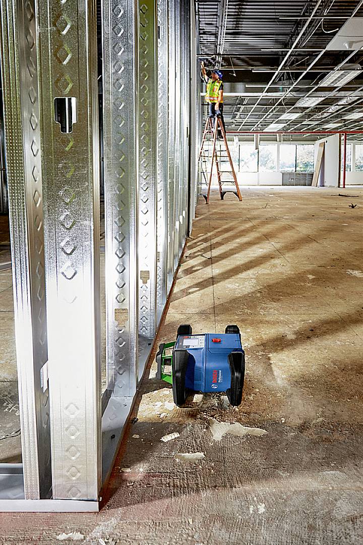 Ein Bauarbeiter auf einer Leiter in einem unfertigen Raum montiert Deckenplatten. Im Vordergrund Metallträger und ein Werkzeugkasten.