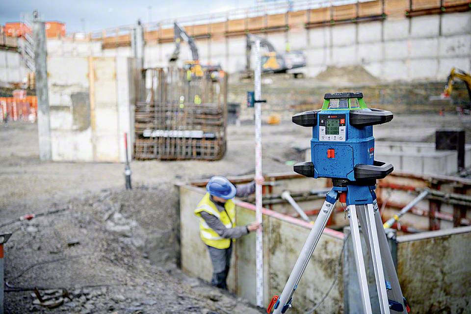 Bauarbeiter vermisst eine Baustelle mit einem Nivelliergerät im Vordergrund; die Umgebung zeigt große Baugruben und Betonwände.