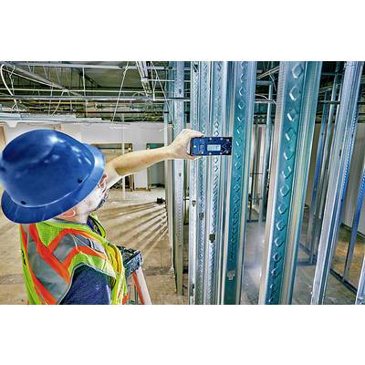 Ein Bauarbeiter in einem neonfarbenen Sicherheitsweste und blauem Helm misst mit einem elektronischen Gerät den Abstand zwischen Metallträgern in einem Rohbau.