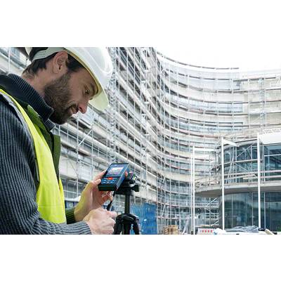 Ein Bauarbeiter in Sicherheitskleidung benutzt ein Vermessungsgerät auf einer Baustelle mit einem großen, im Bau befindlichen Gebäude.