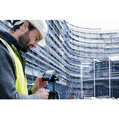 Ein Bauarbeiter mit Sicherheitshelm und Weste verwendet ein Laser-Messgerät auf einer Baustelle vor einem eingerüsteten Gebäude.