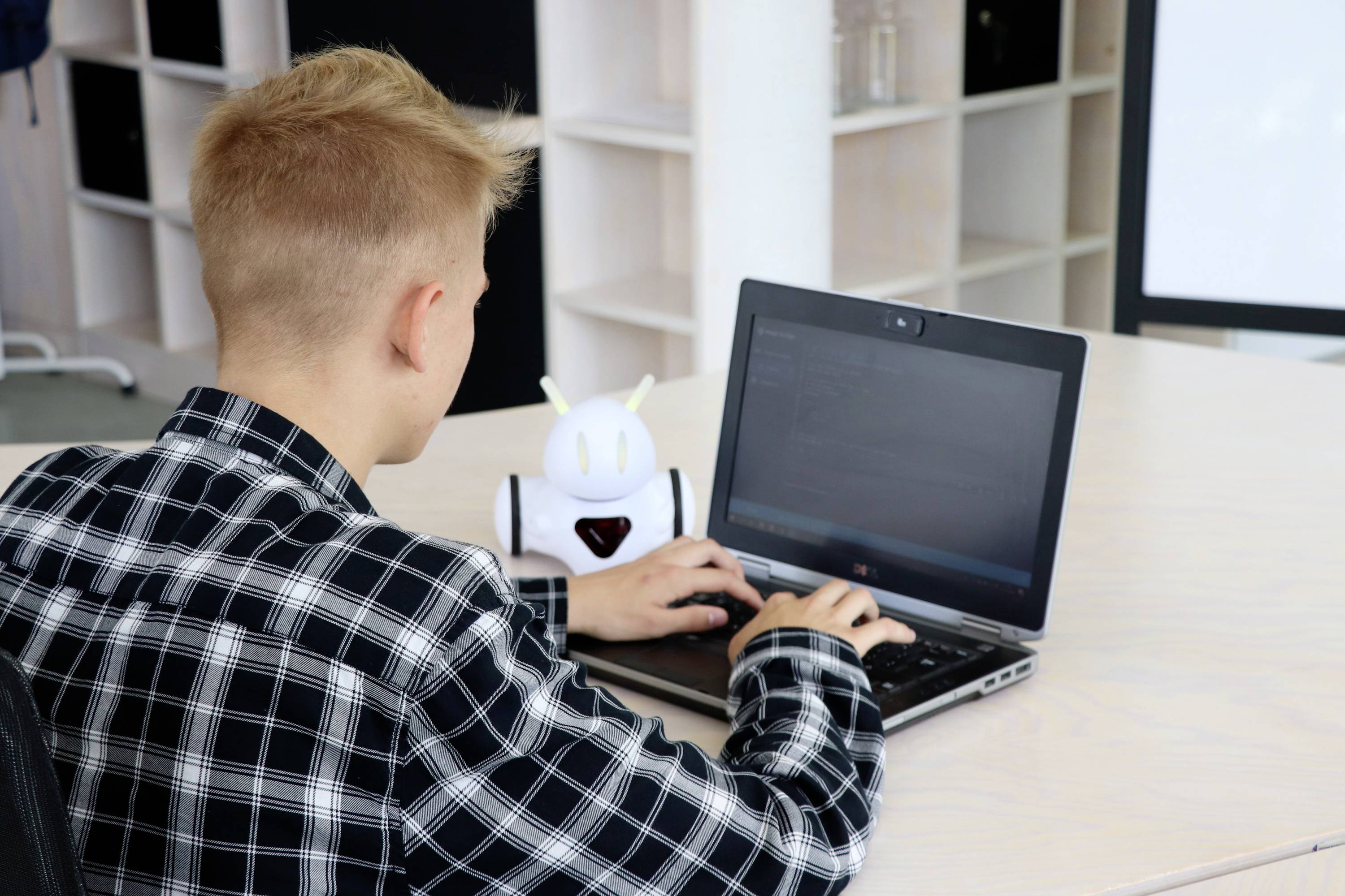 Ein junger Mensch arbeitet am Laptop in einem modernen Büro. Neben ihm steht ein kleiner Roboter. Im Hintergrund sind Regale sichtbar.
