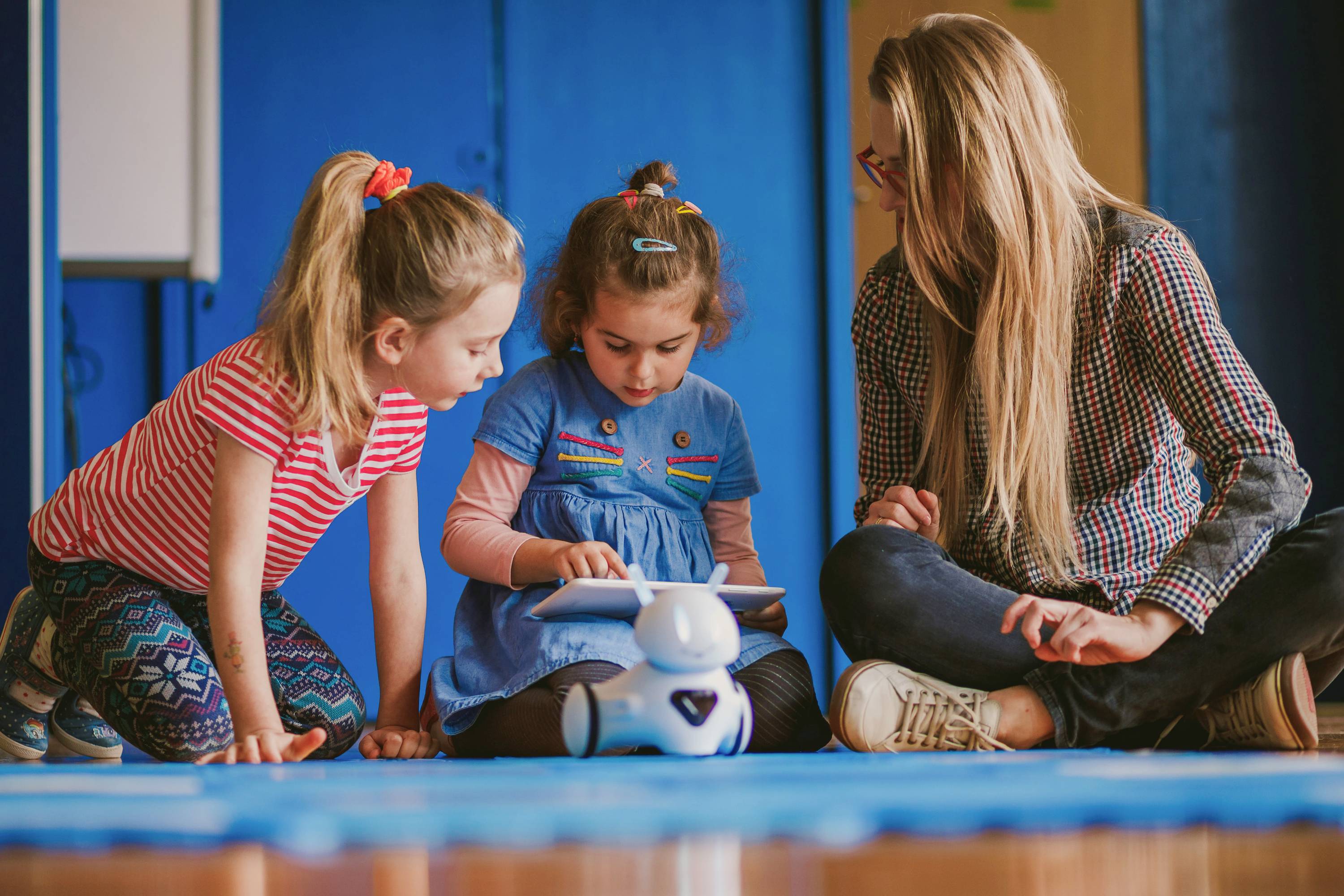 Drei Kinder sitzen auf dem Boden und spielen mit einem kleinen weißen Roboter, der einem Hund ähnelt. Eine Frau beobachtet und lächelt.