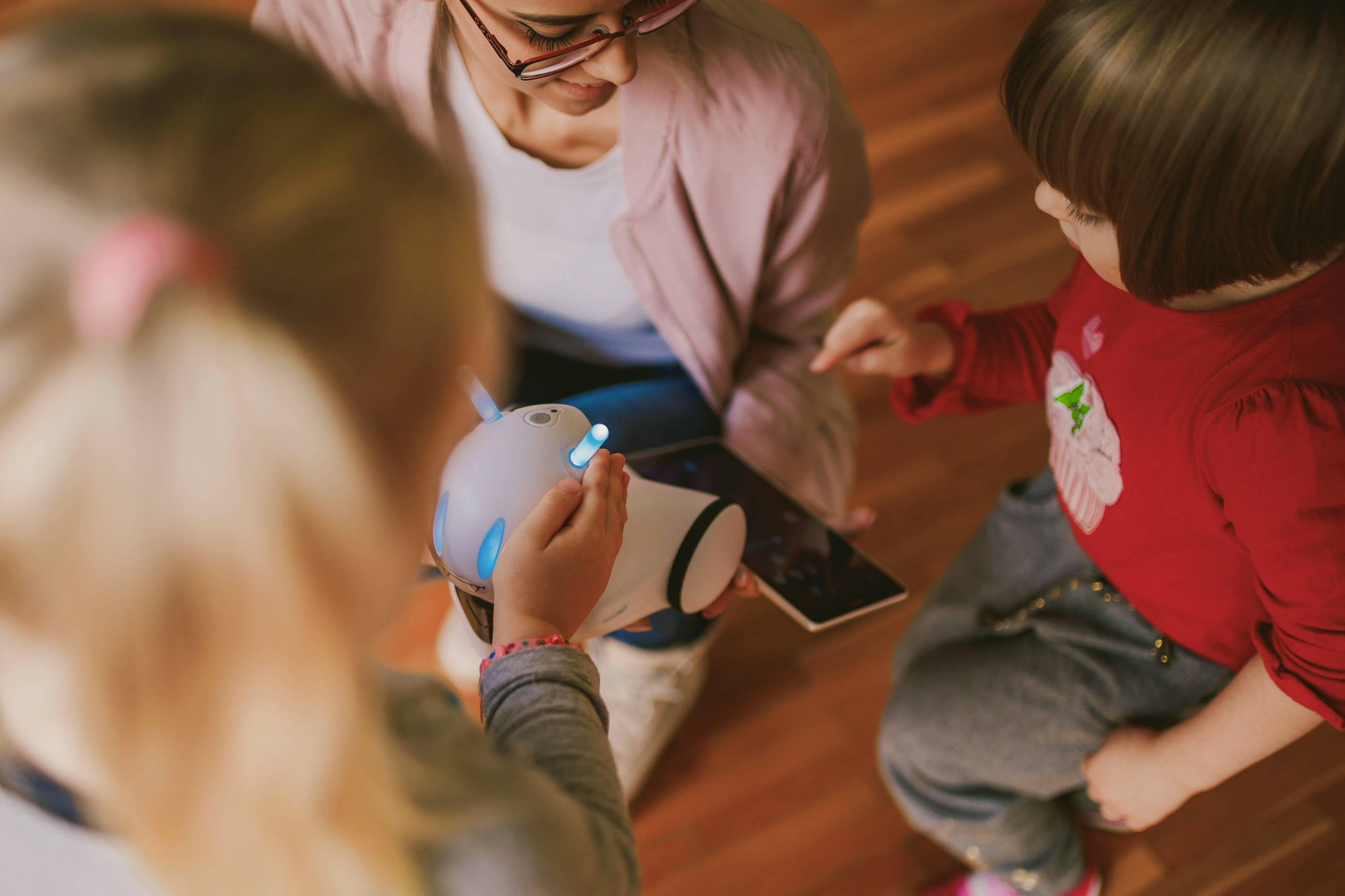 Eine erwachsene Person und zwei Kinder spielen mit einem kleinen Roboter auf einem Holzboden. Ein Tablet liegt daneben.