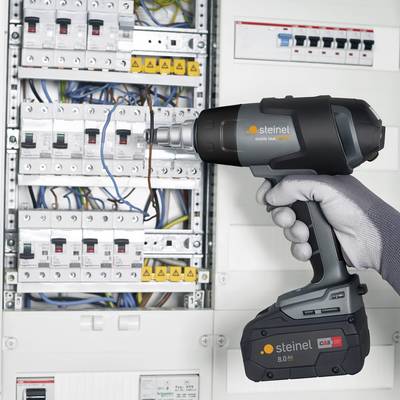 Ein Handwerker hält ein Profi-Heißluftgebläse vor einem geöffneten Schaltschrank mit zahlreichen Kabeln und Schaltern.