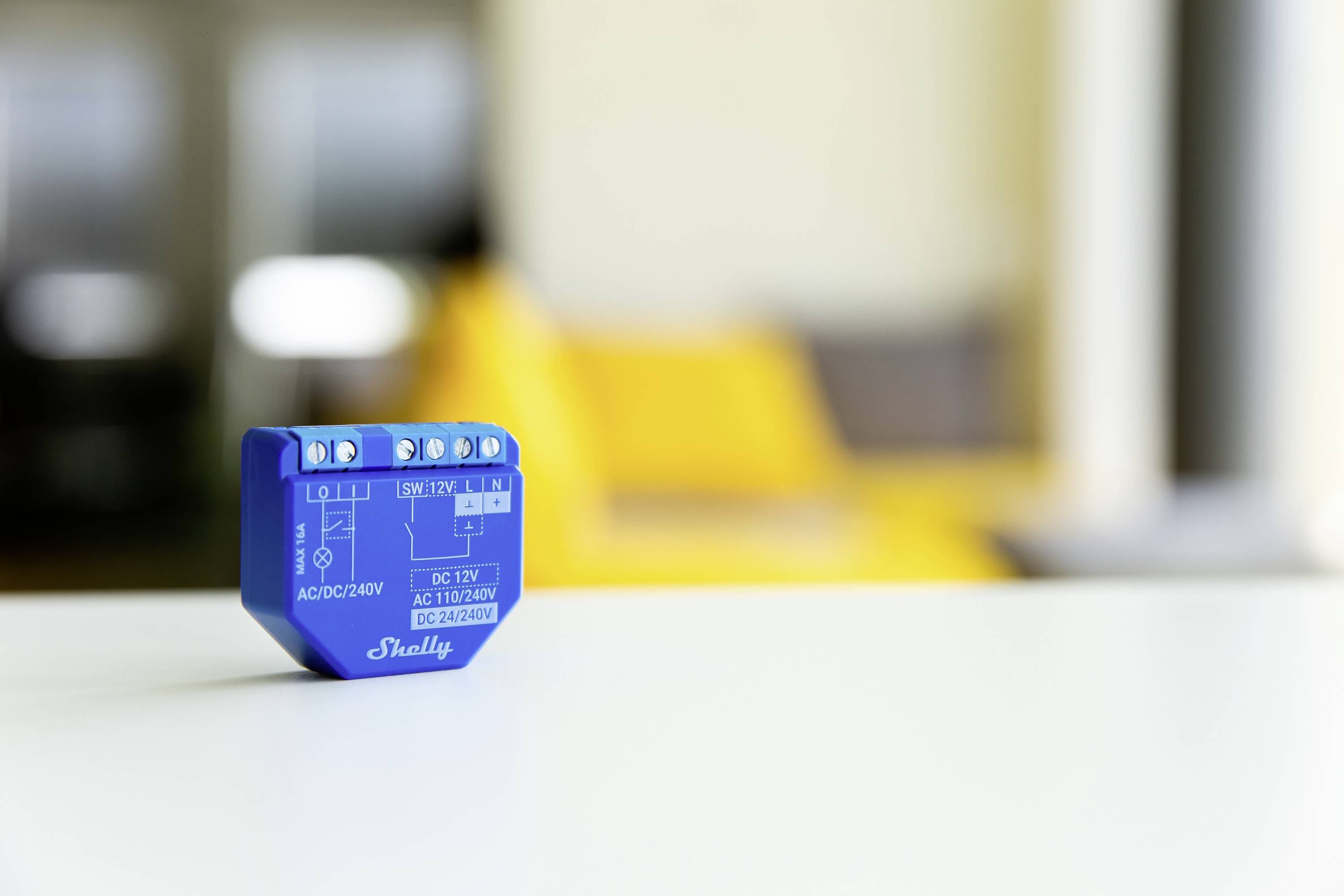 Ein blaues elektronisches Bauteil sitzt auf einem weißen Tisch vor einem gelben Sofa. Es scheint ein Relais oder Schalter zu sein.