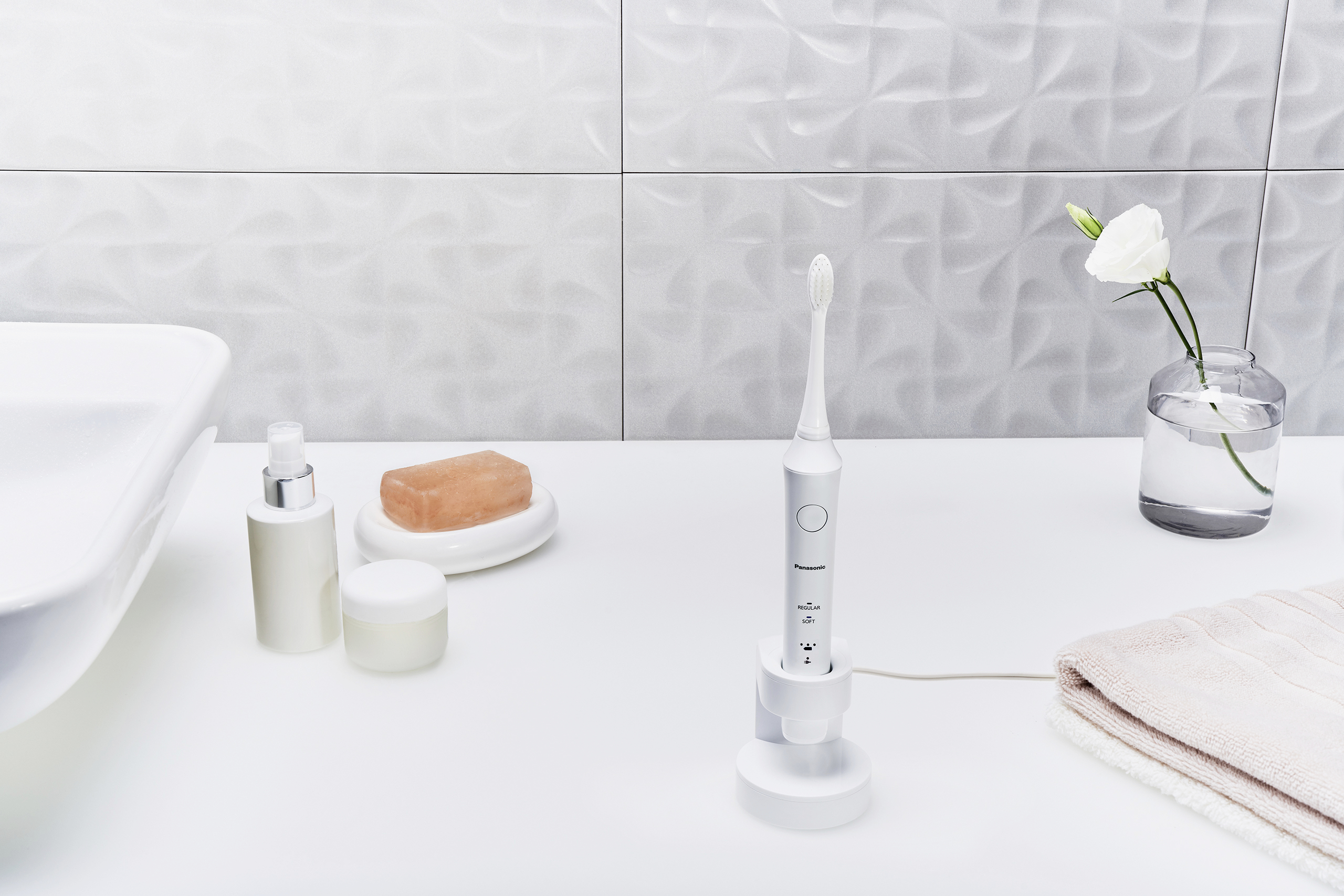 Elektrische Zahnbürste auf weißem Badezimmer-Waschbecken, neben Handtuch, Seife und Vase mit weißer Blume.