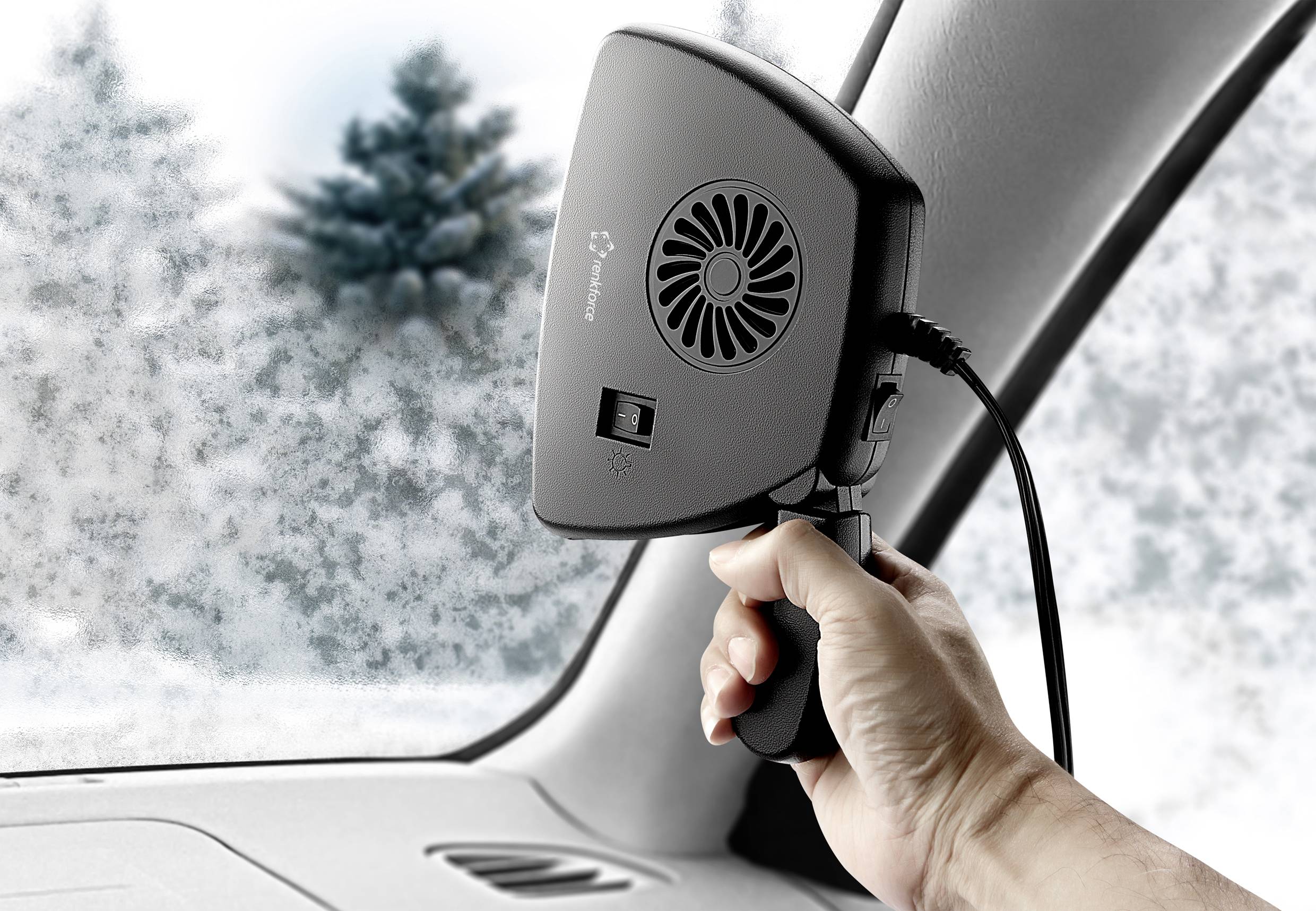 Eine Hand hält einen tragbaren Auto-Heizlüfter nahe der Windschutzscheibe. Draußen sind verschneite Bäume sichtbar.