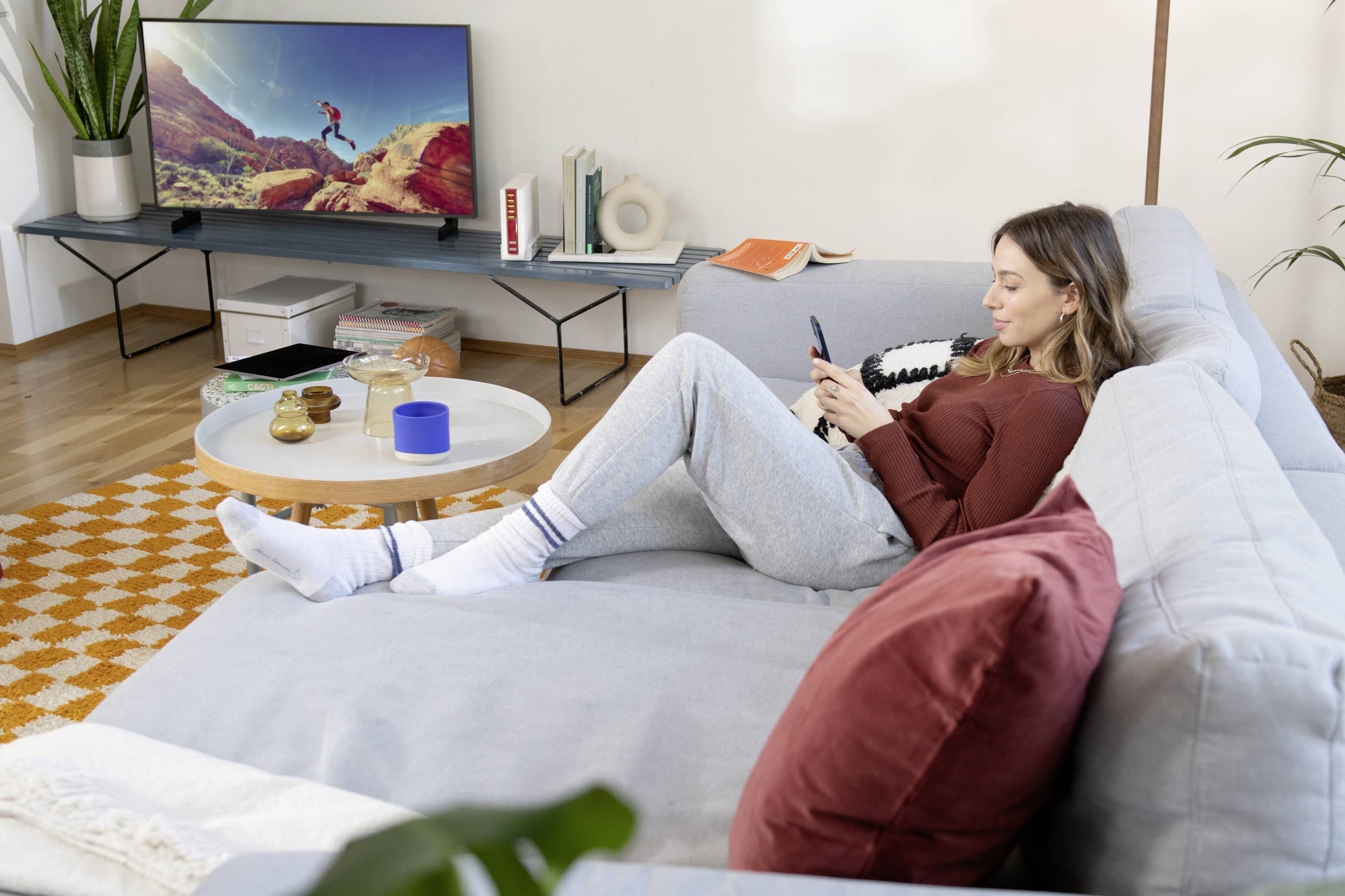 Eine Person sitzt entspannt auf einem Sofa und schaut auf ihr Handy. Im Hintergrund ist ein Fernseher eingeschaltet, der eine Landschaft zeigt.