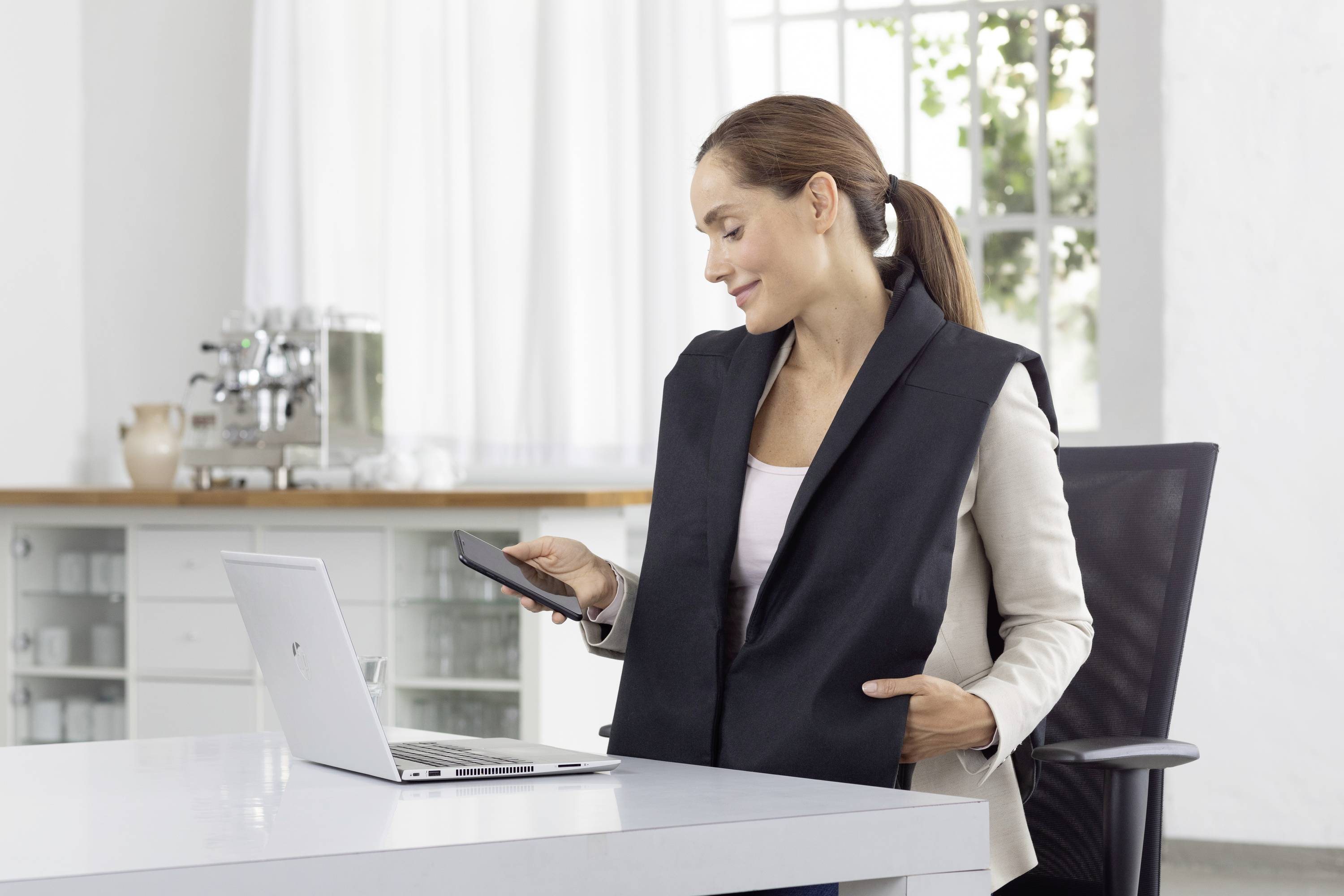 Eine Frau sitzt an einem Schreibtisch mit einem Laptop, hält ein Smartphone in der Hand und trägt ein schwarzes Schulterkissen.