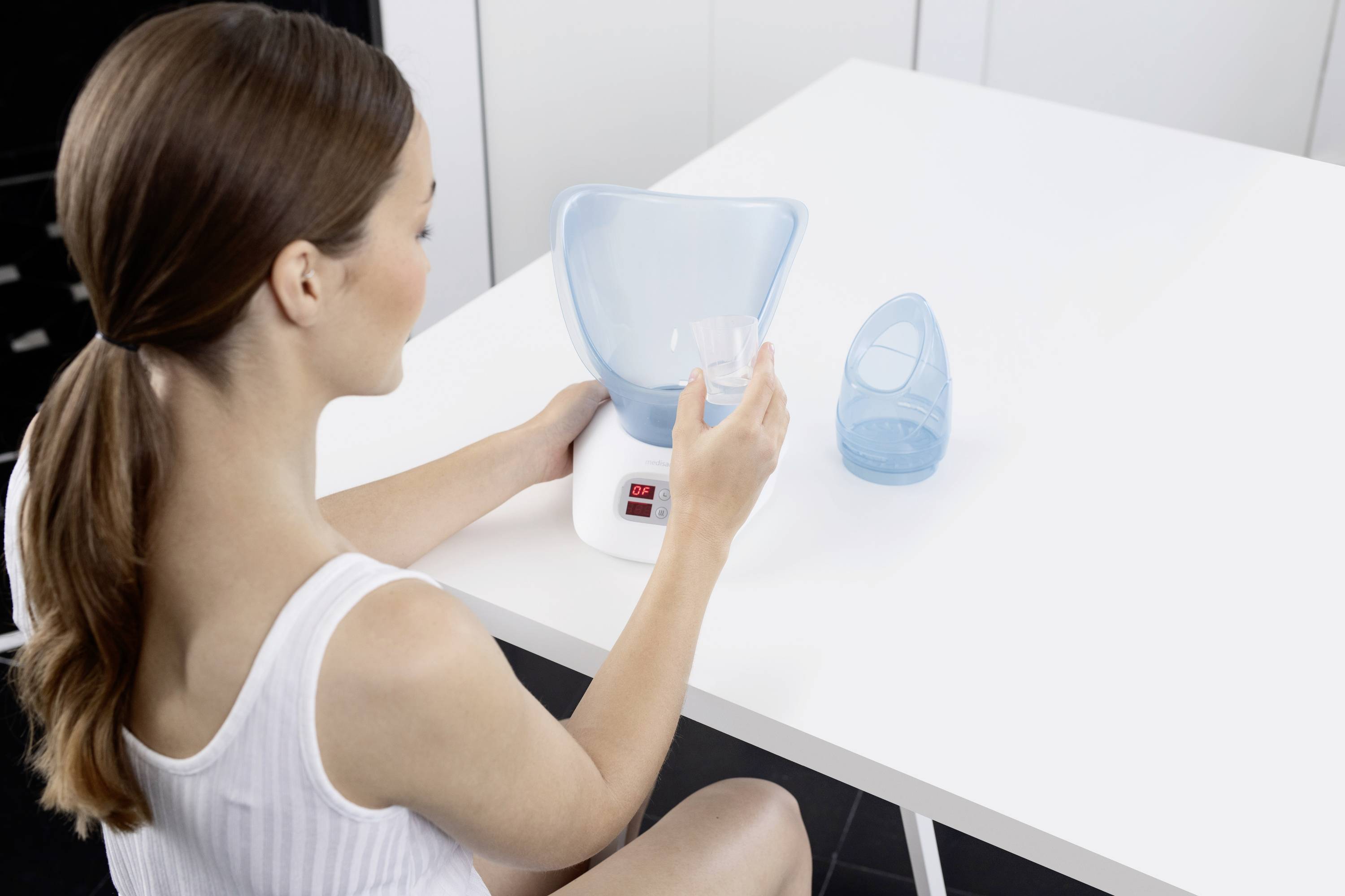 Eine Frau benutzt einen Gesichtsinhalator auf einem Tisch. Der Inhalator ist weiß mit einem blauen Aufsatz.