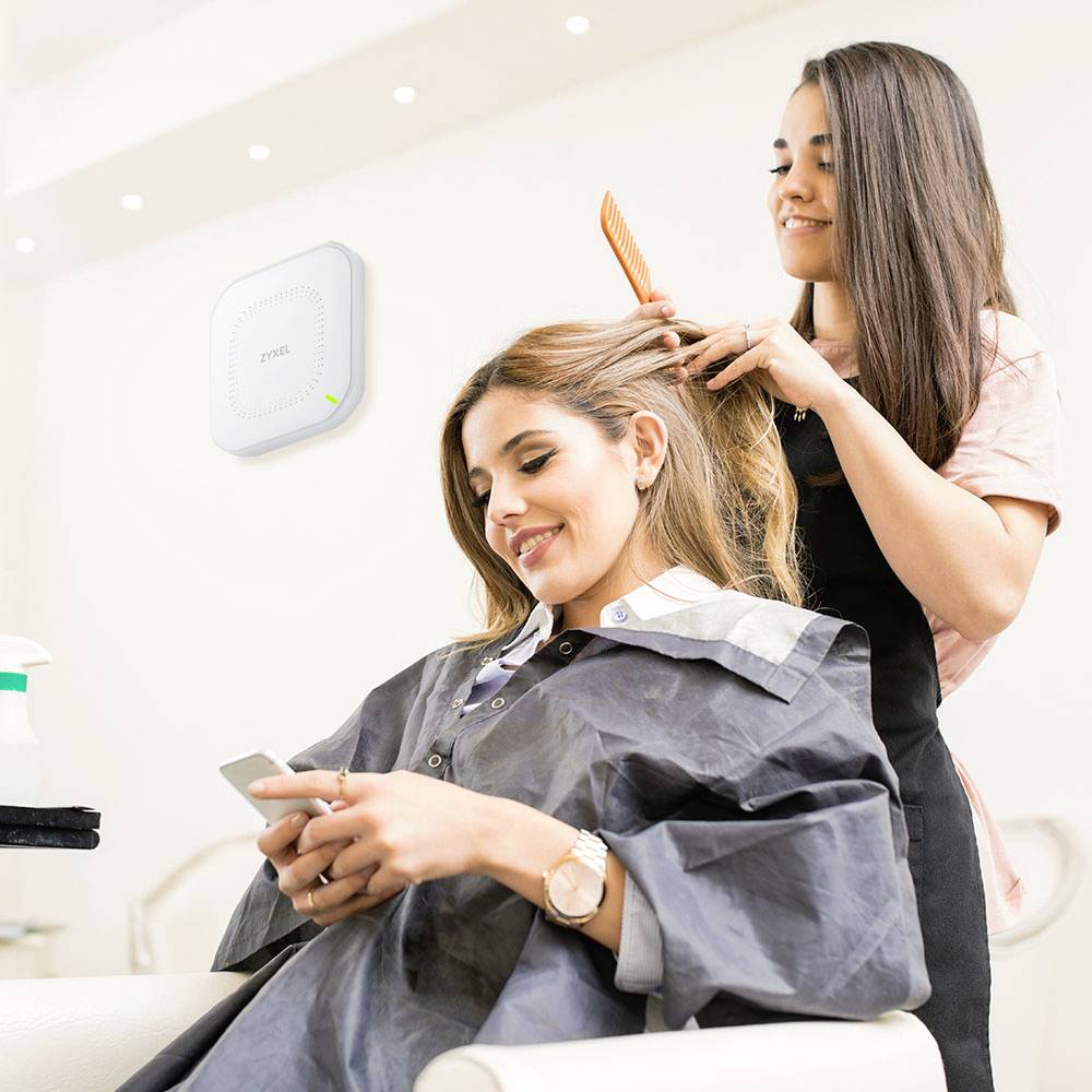 Eine Friseurin stylt mit einem Kamm das Haar einer sitzenden Frau, die auf ihr Handy schaut. Im Hintergrund ist ein WLAN-Router an der Wand.