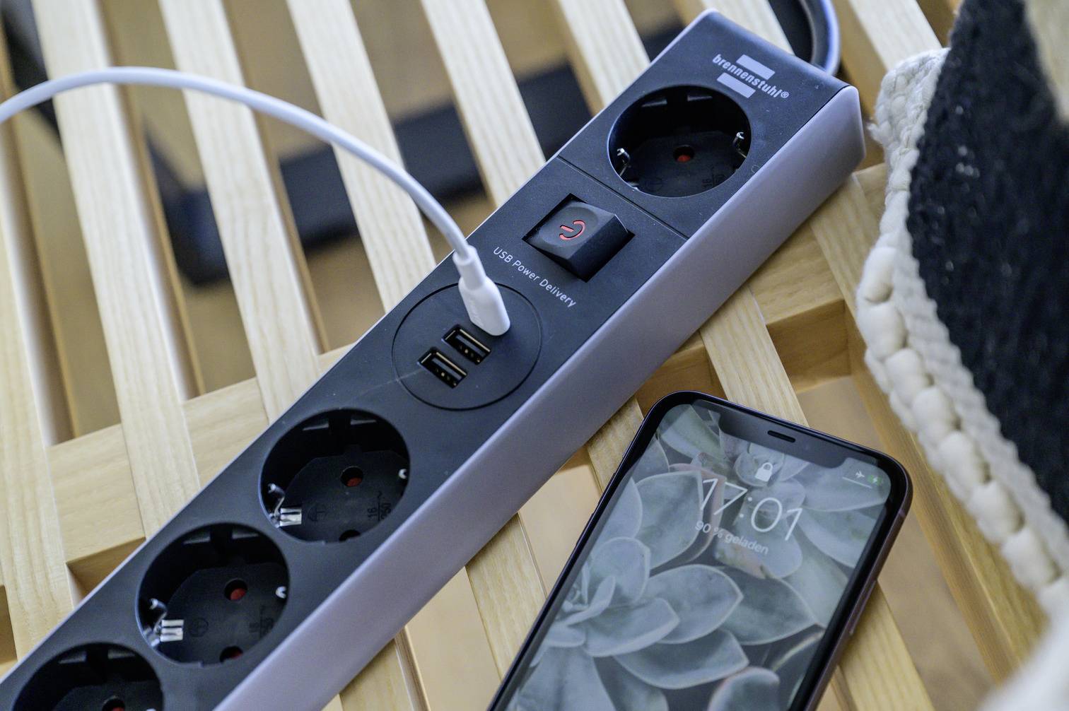 Eine Mehrfachsteckdose mit USB-Anschluss liegt auf einem Holztisch. Ein Kabel ist eingesteckt. Daneben liegt ein Smartphone mit einer Pflanzen-Anzeige.