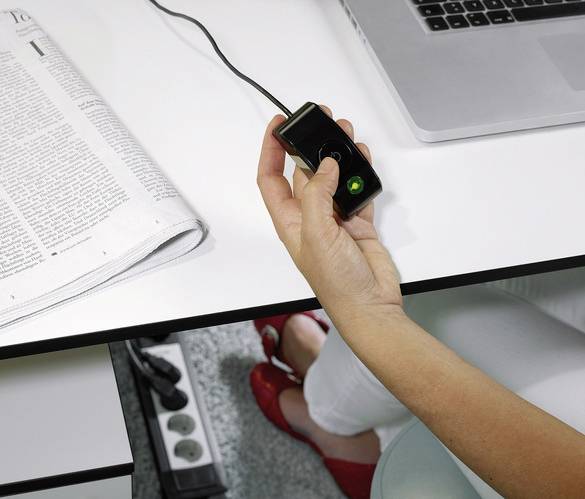 Eine Person hält einen kabelgebundenen Fingerabdruckscanner neben einem Laptop und einer Zeitung. Im Hintergrund sind rote Schuhe zu sehen.
