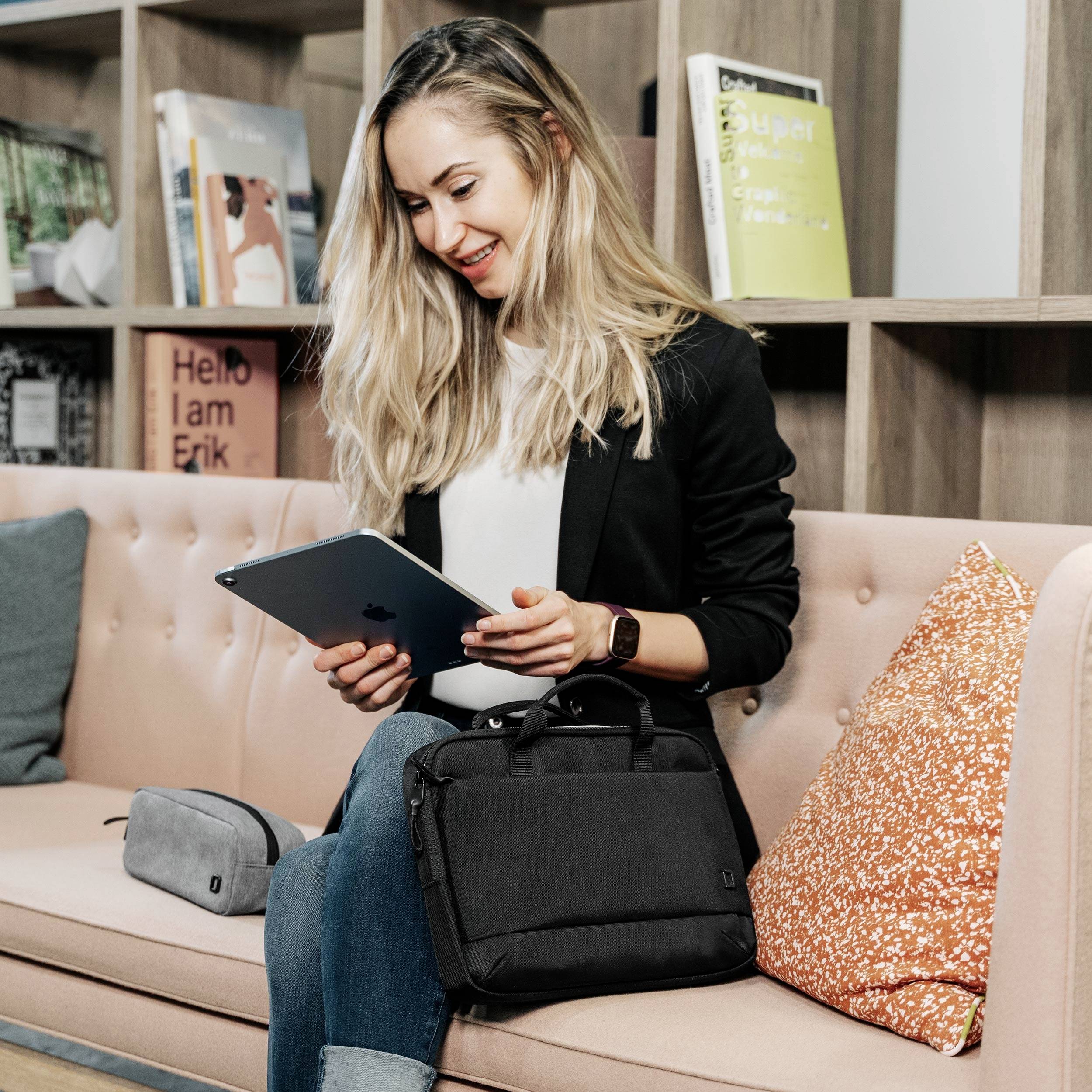 Eine Frau sitzt auf einem Sofa, schaut lächelnd auf ein Tablet. Neben ihr eine Tasche und im Hintergrund ein Bücherregal.