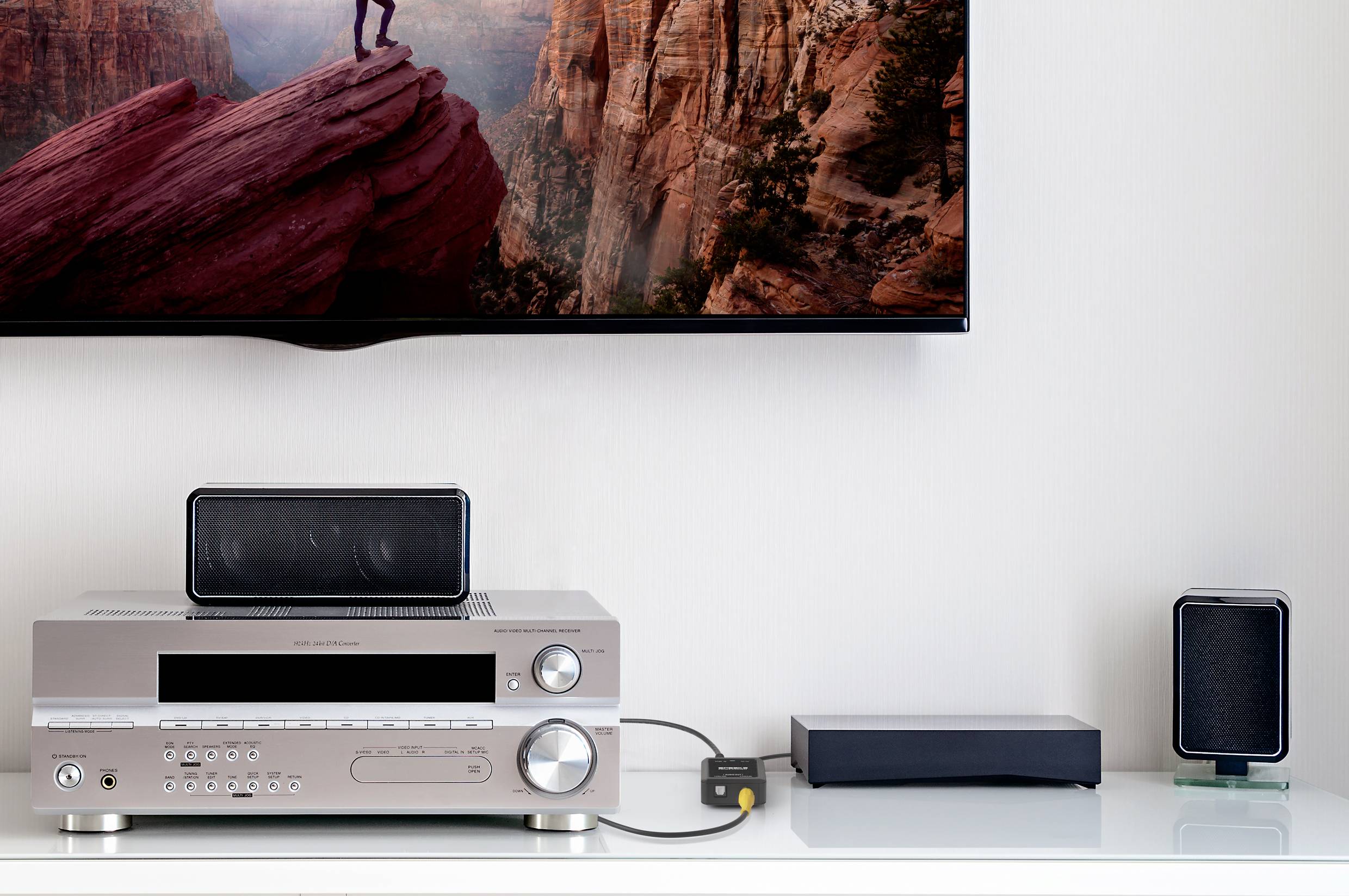 Ein modernes Wohnzimmer mit einem TV an der Wand, einem Verstärker, zwei Lautsprechern und einer Set-Top-Box auf einem Regal darunter.