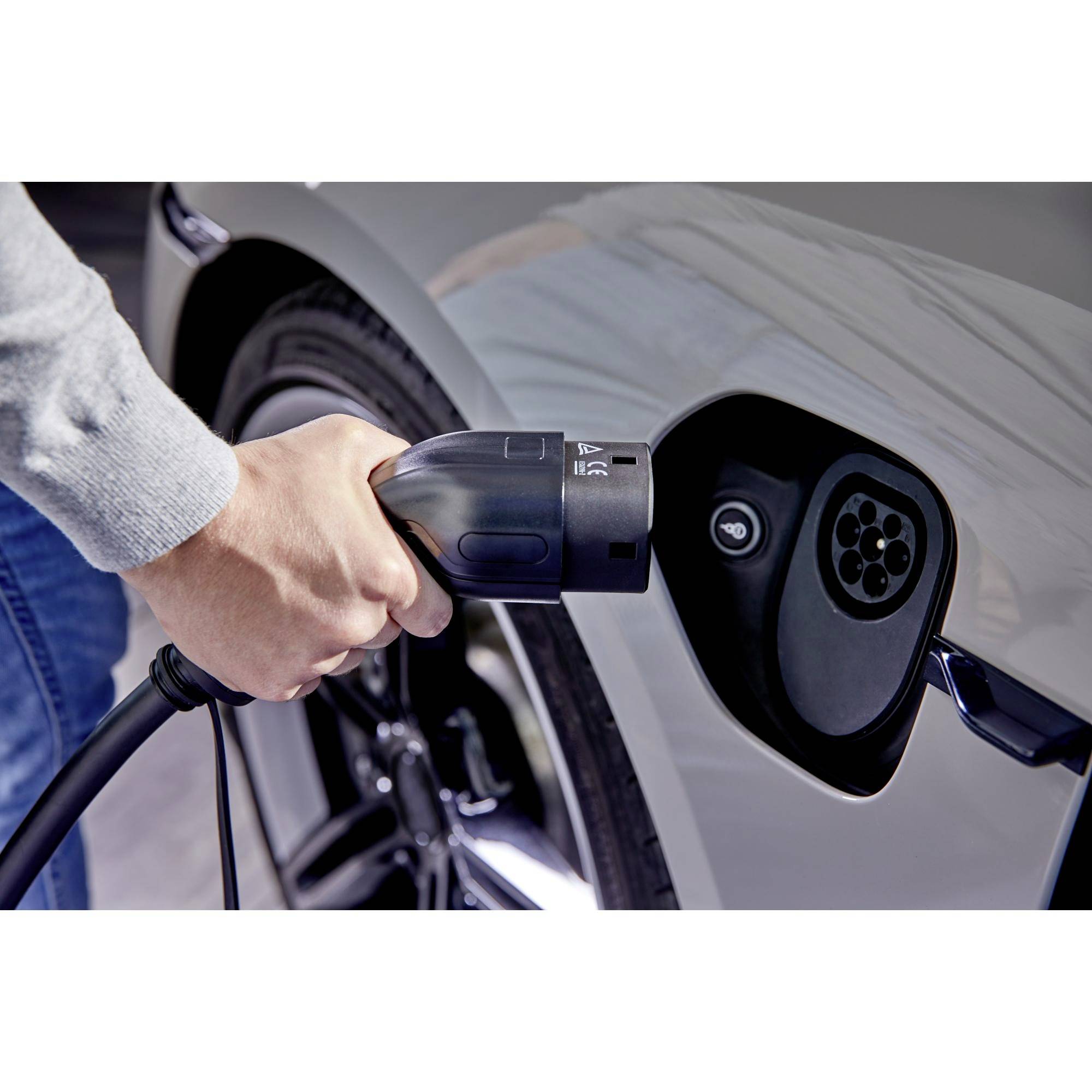Eine Hand hält einen schwarzen Ladestecker und verbindet ihn mit einem Elektroauto, das an einer Ladestation angeschlossen wird.