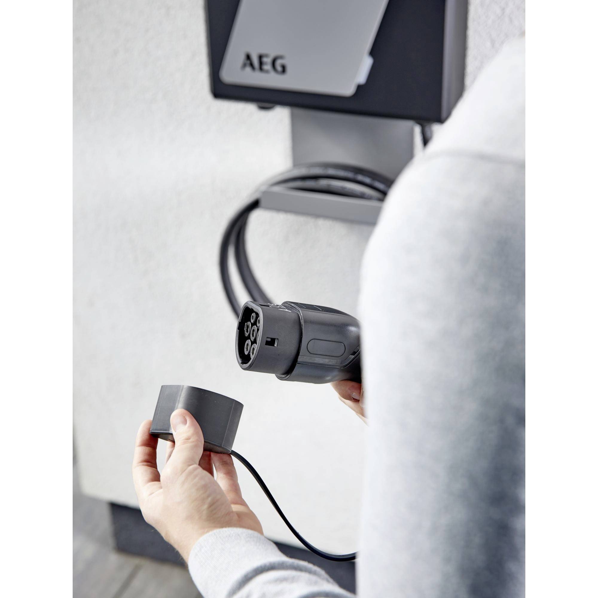 Eine Person hält ein Ladekabel für ein Elektrofahrzeug vor einer Ladestation an der Wand.