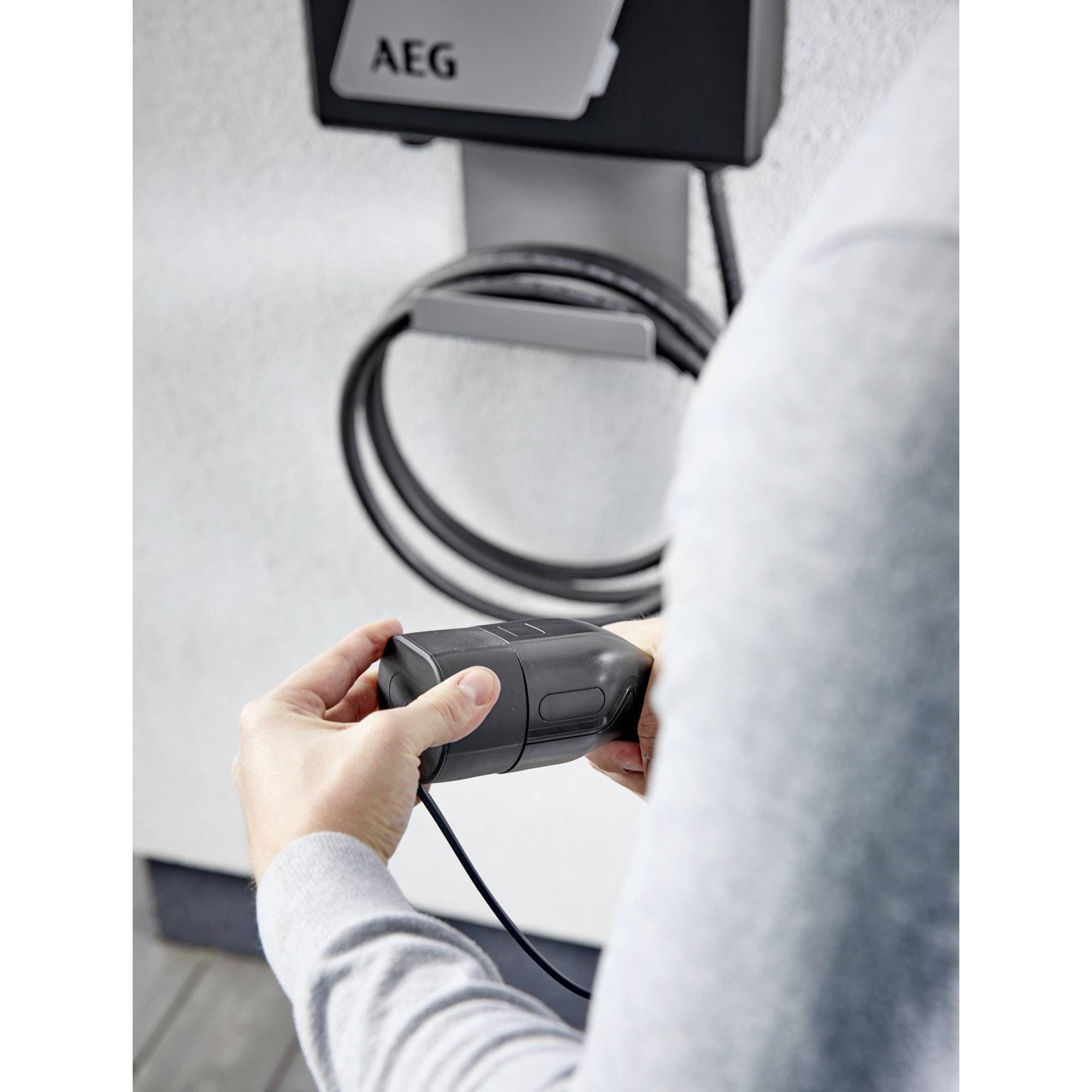 Eine Person hält ein E-Ladekabel vor einer AEG-Wandladestation. Das Kabel ist bereit zum Anschließen an ein Elektrofahrzeug.