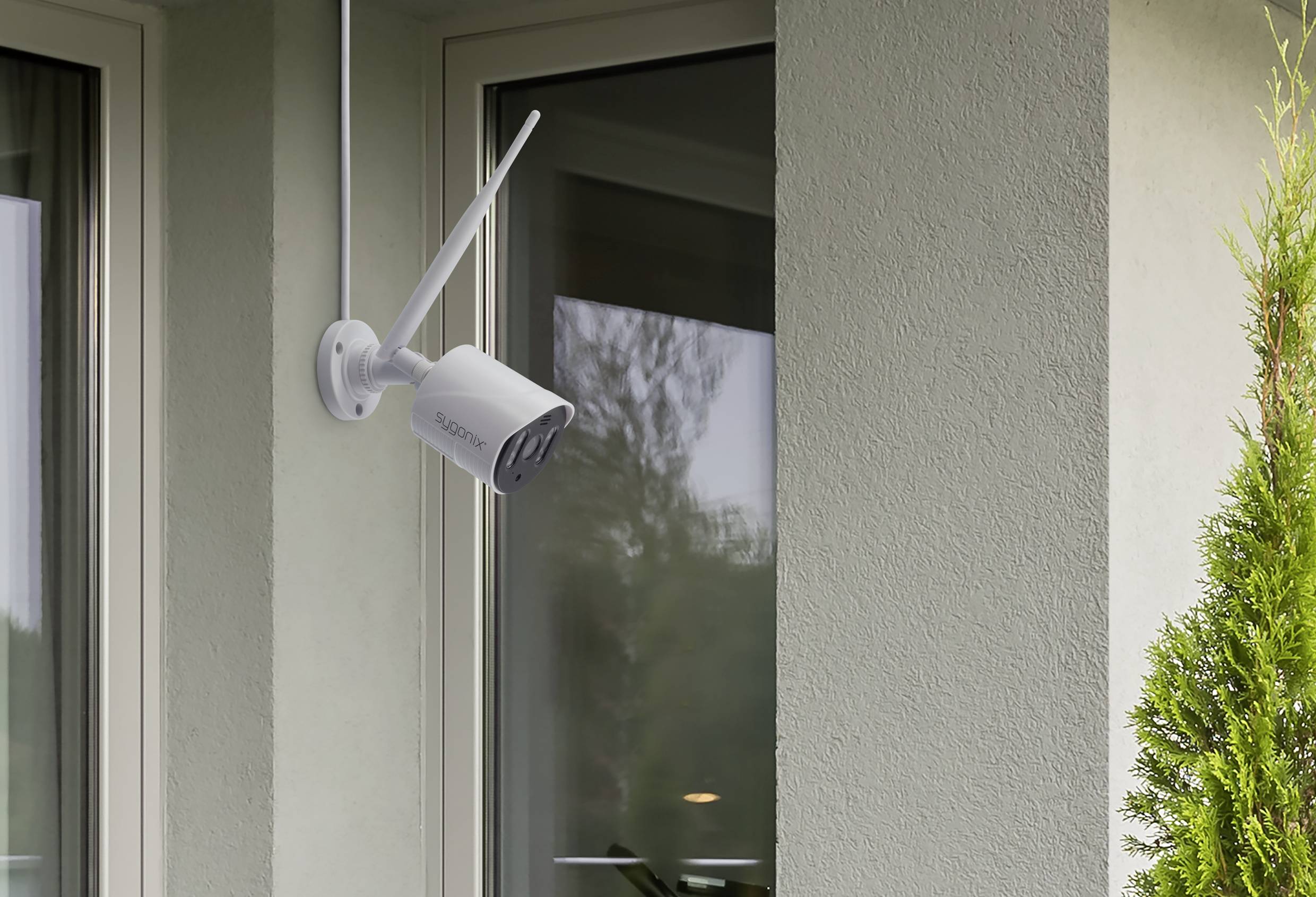 Überwachungskamera an einer Außenecke eines Gebäudes installiert, mit Blick auf Fenster und umgeben von grüner Vegetation.