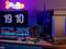 Gaming-Setup mit Monitor, PC-Gehäuse, beleuchteter Tastatur und Headset. Im Hintergrund LED-Lichter und eine Uhranzeige '19:10'.