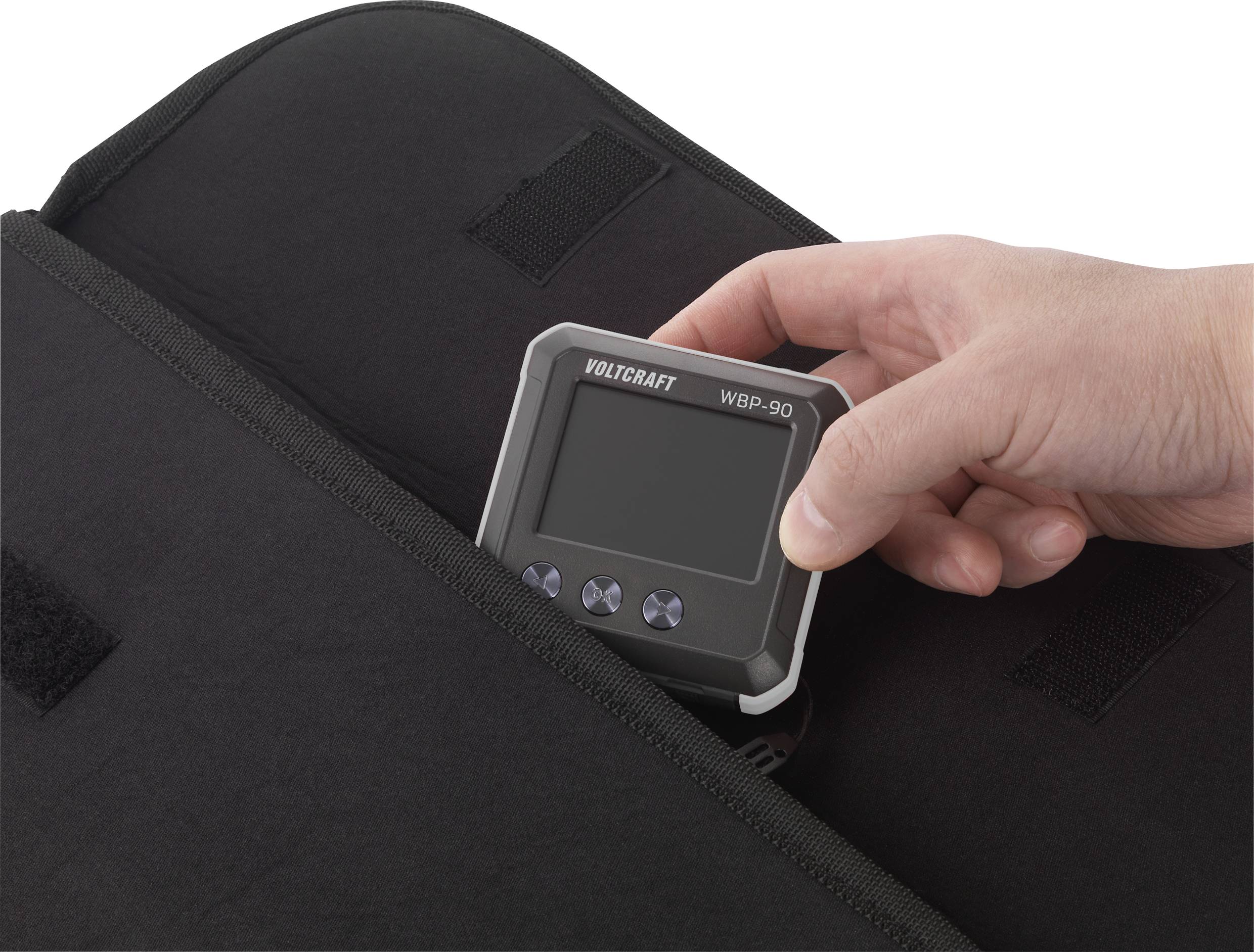 Eine Hand legt ein digitales Gerät, ein Volcraft WBP-90, in eine schwarze Tasche. Das Gerät hat ein digitales Display und Tasten.