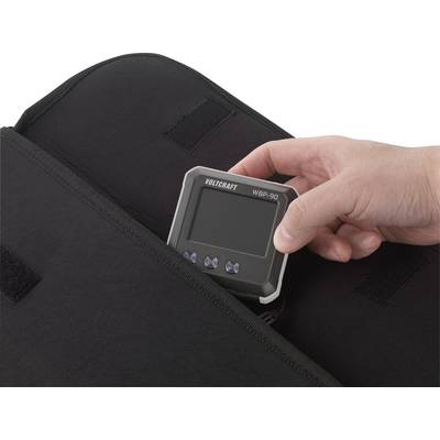 Eine Hand legt ein digitales Gerät, ein Volcraft WBP-90, in eine schwarze Tasche. Das Gerät hat ein digitales Display und Tasten.