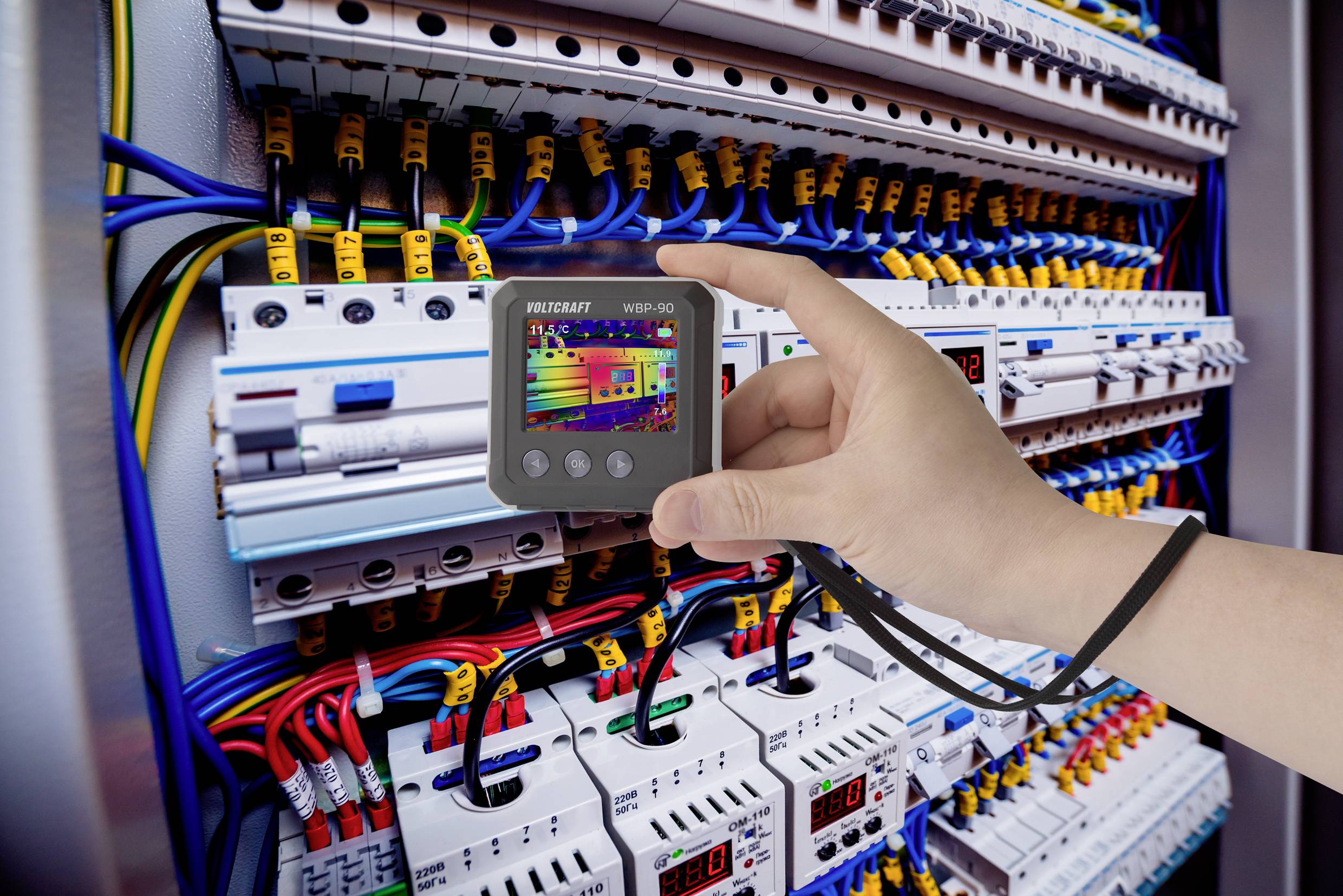 Eine Hand hält ein Prüfgerät vor einen geöffneten Schaltschrank mit sichtbarer Verkabelung, um elektrische Messungen durchzuführen.