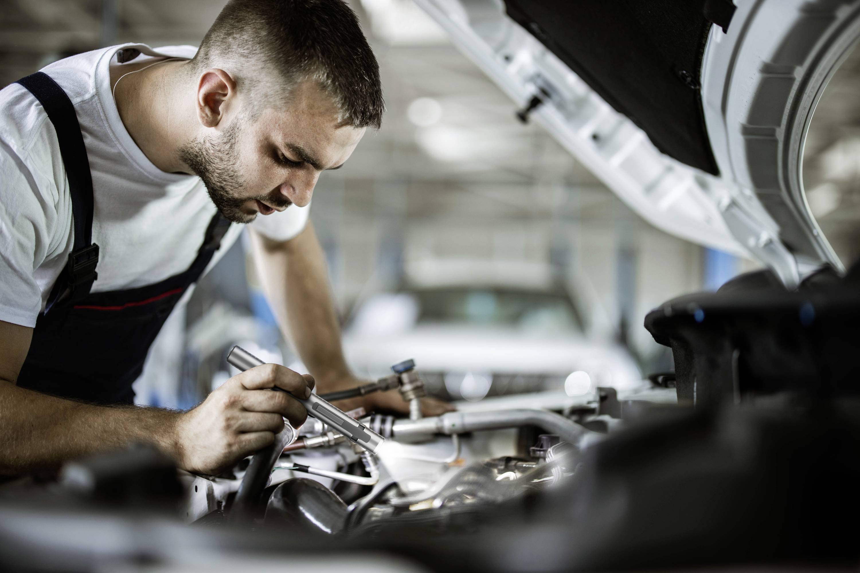 Ein Mechaniker inspiziert mit Werkzeug den Motorraum eines Autos in einer Werkstattumgebung.