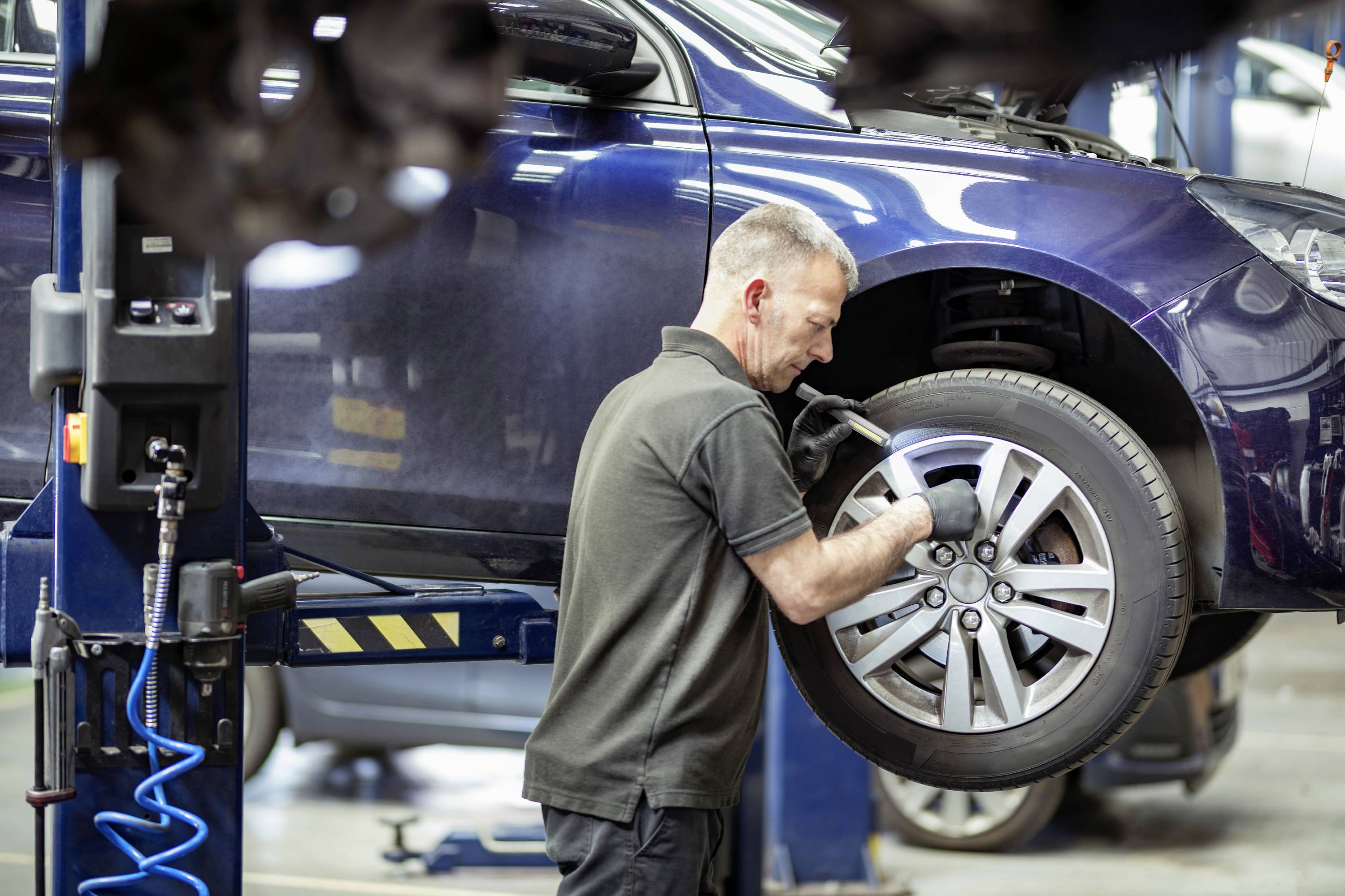 Ein Mechaniker überprüft den Reifen eines angehobenen blauen Autos in einer Werkstatt.