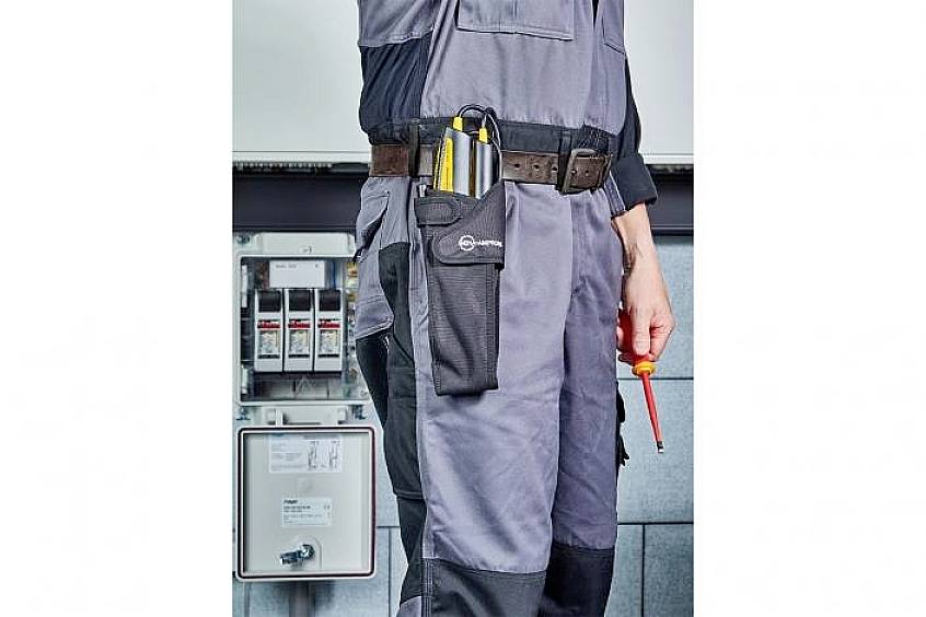 Ein Elektriker trägt ein graues Arbeitsoutfit und hält einen Schraubenzieher. Ein Multimeter ist in einer Tasche an seinem Gürtel. Im Hintergrund sind Sicherungskästen zu sehen.