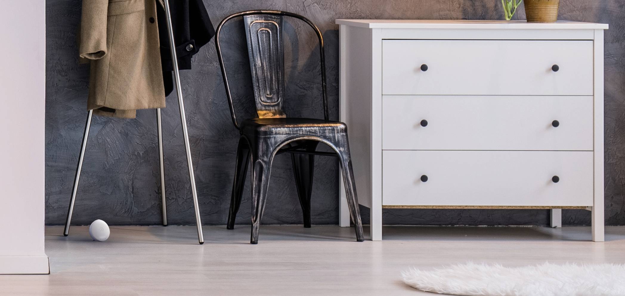 Ein Metallsessel steht zwischen einem hellen Schrank mit drei Schubladen und einem beigen Mantel, der an einem Kleiderständer hängt.