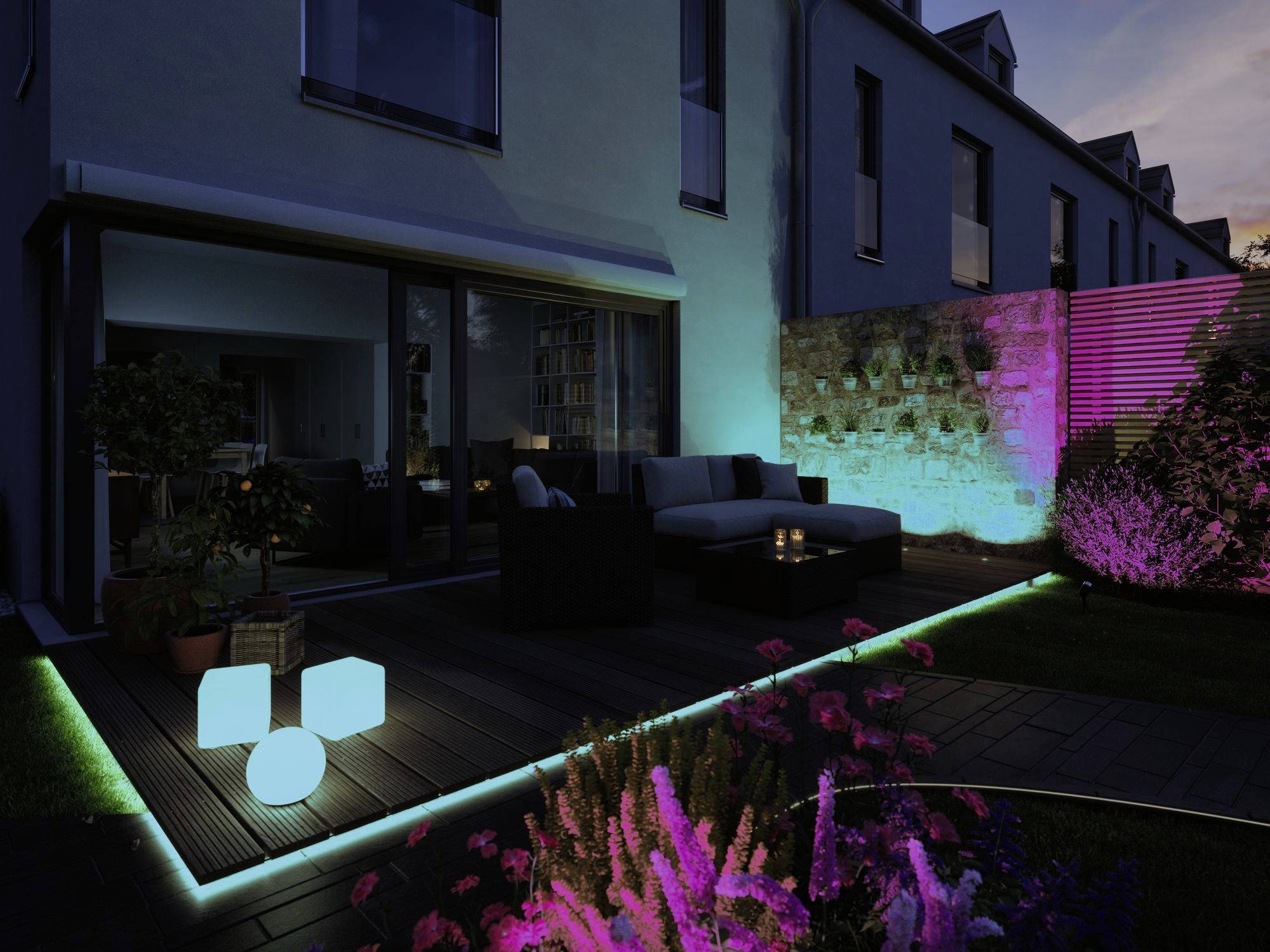 Ein Garten bei Dämmerung mit bunten LED-Lichtern: leuchtende Kuben auf dem Boden, in den Pflanzen und entlang der Terrassenkante.