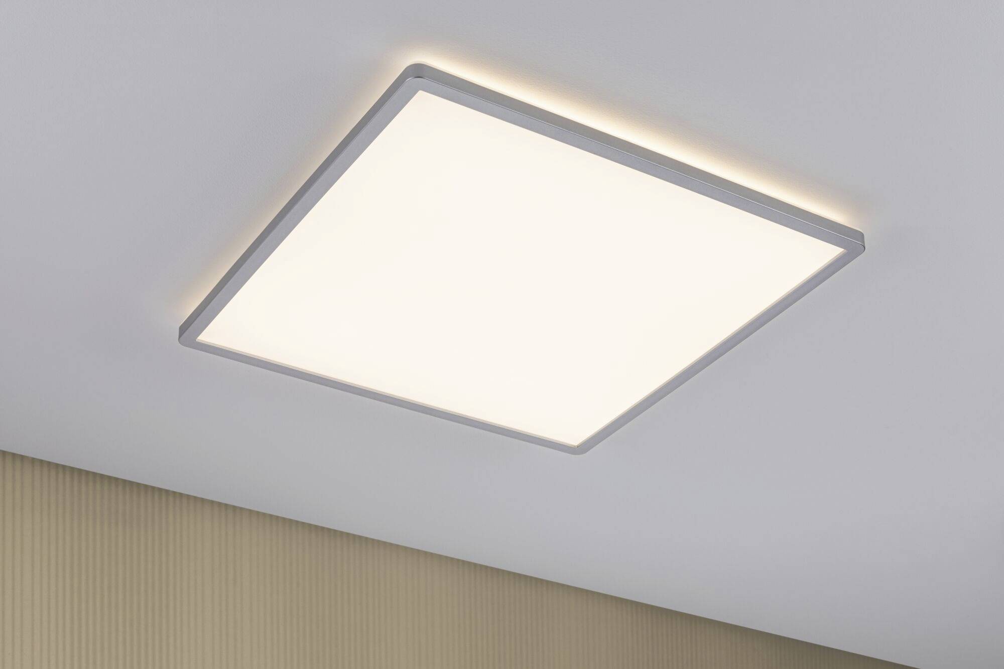 Eine quadratische Deckenleuchte, die sanftes Licht in einem Raum verteilt. Die Decke ist weiß, die Wand hat eine leicht gestreifte Textur.