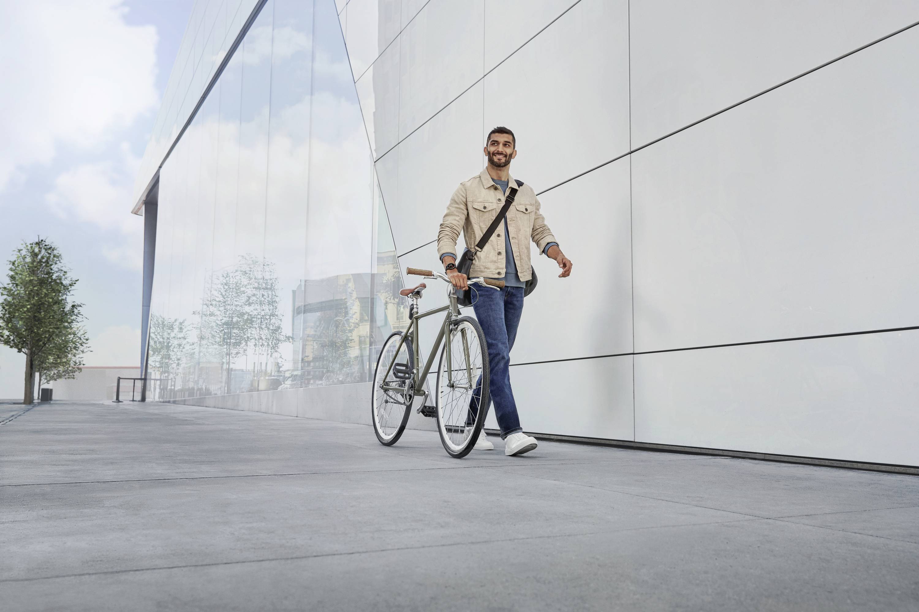 Ein Mann geht neben einem Fahrrad entlang eines modernen Gebäudes auf einem Gehweg bei sonnigem Wetter.