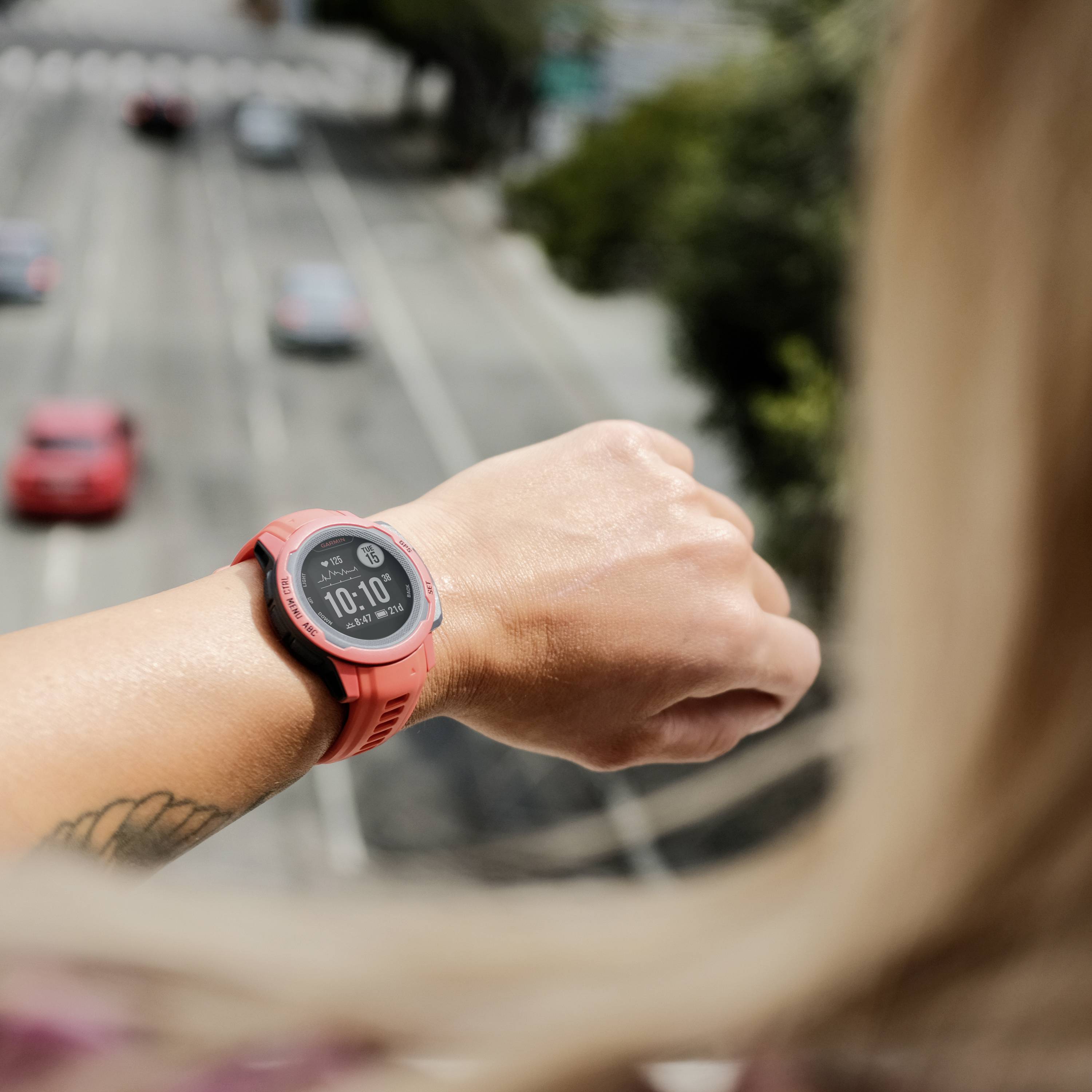Eine Person mit einer Sportuhr schaut auf die Uhrzeit auf einer Brücke über einer mehrspurigen Straße mit vorbeifahrenden Autos.
