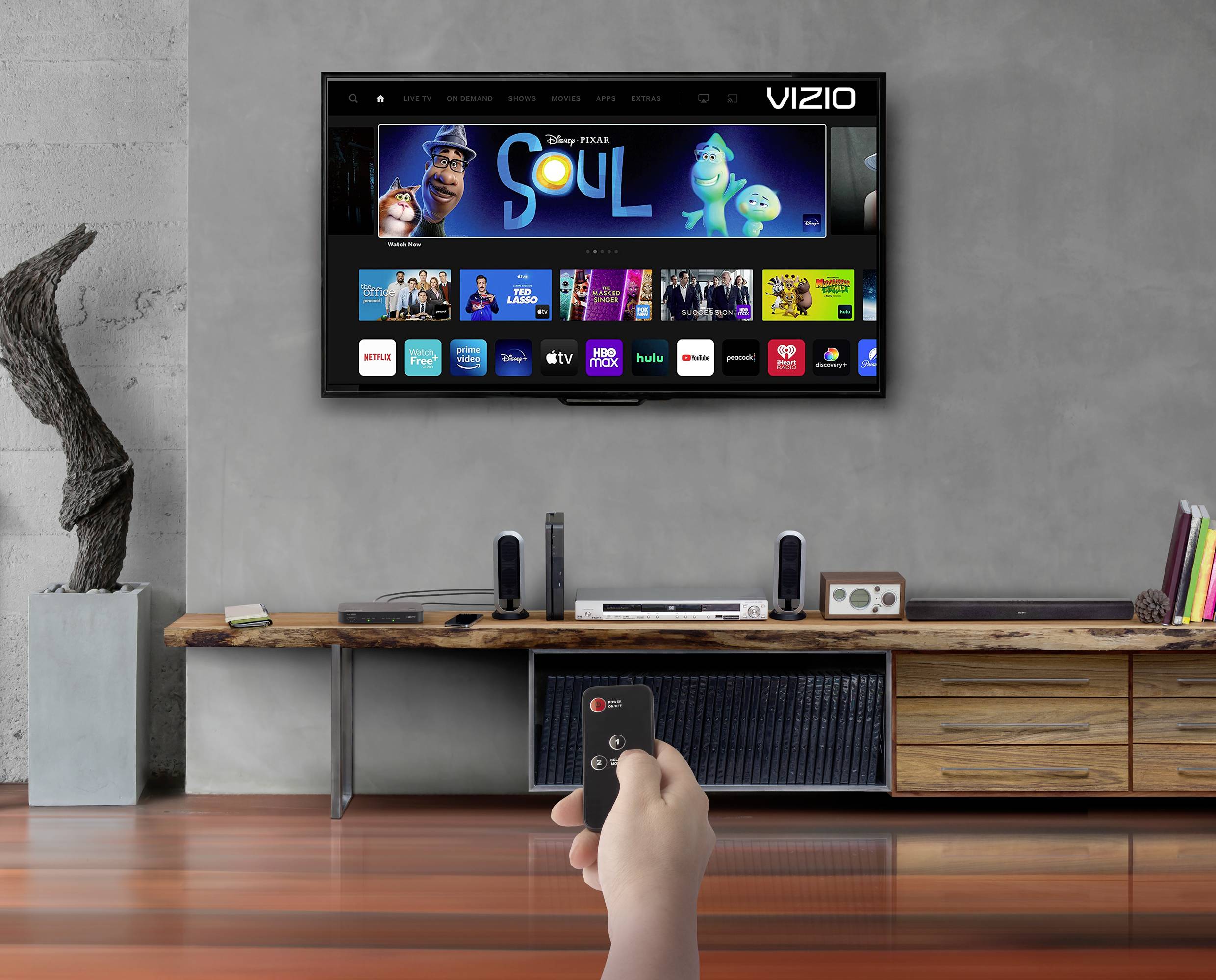 Ein Wohnzimmer mit moderner Einrichtung. Ein Fernseher an der Wand zeigt das Menü eines Streaming-Dienstes. Jemand hält eine Fernbedienung.