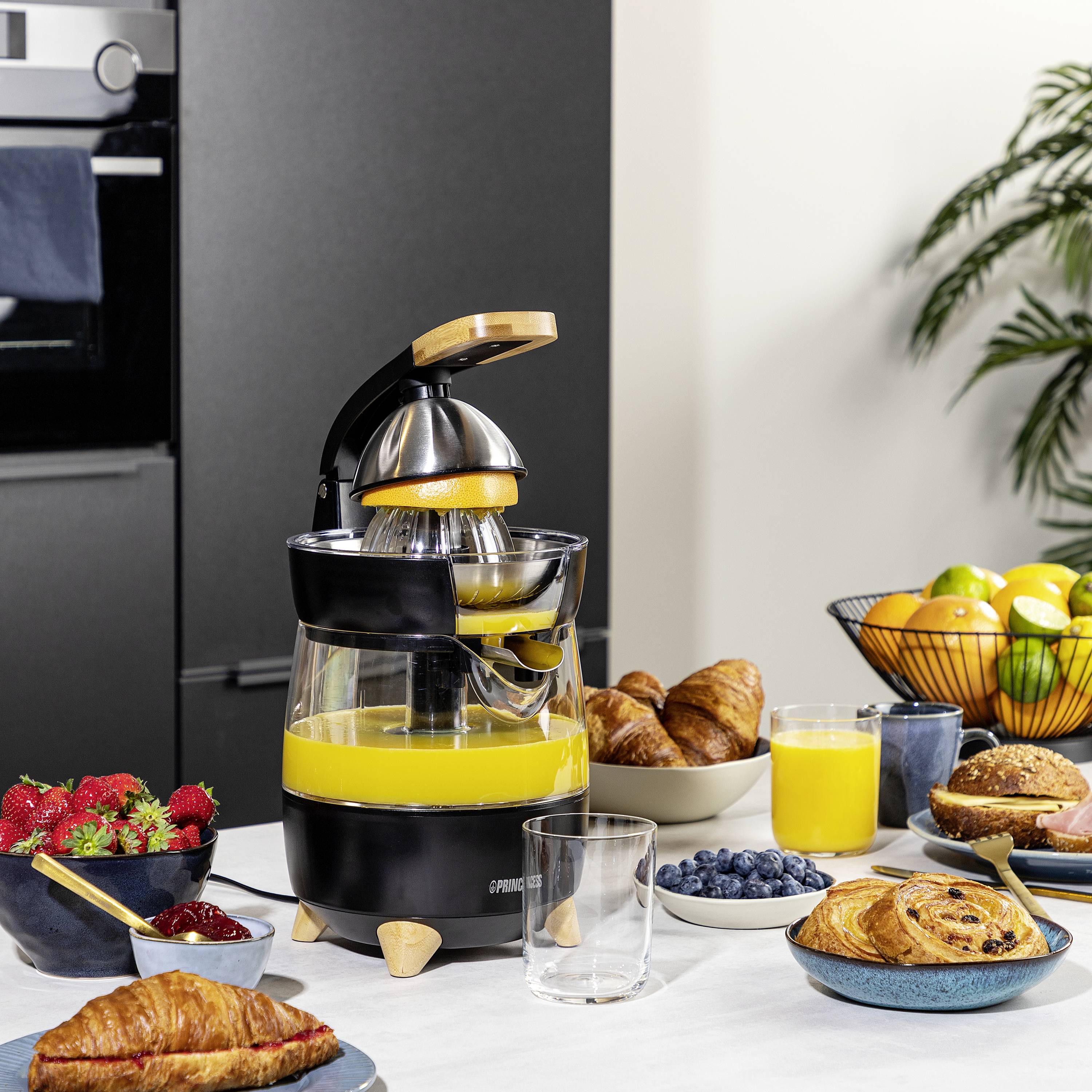 Eine Saftpresse aus Glas mit frischem Orangensaft steht auf einem Tisch. Daneben sind Croissants, Obstschale, Beeren und ein Glas Saft.