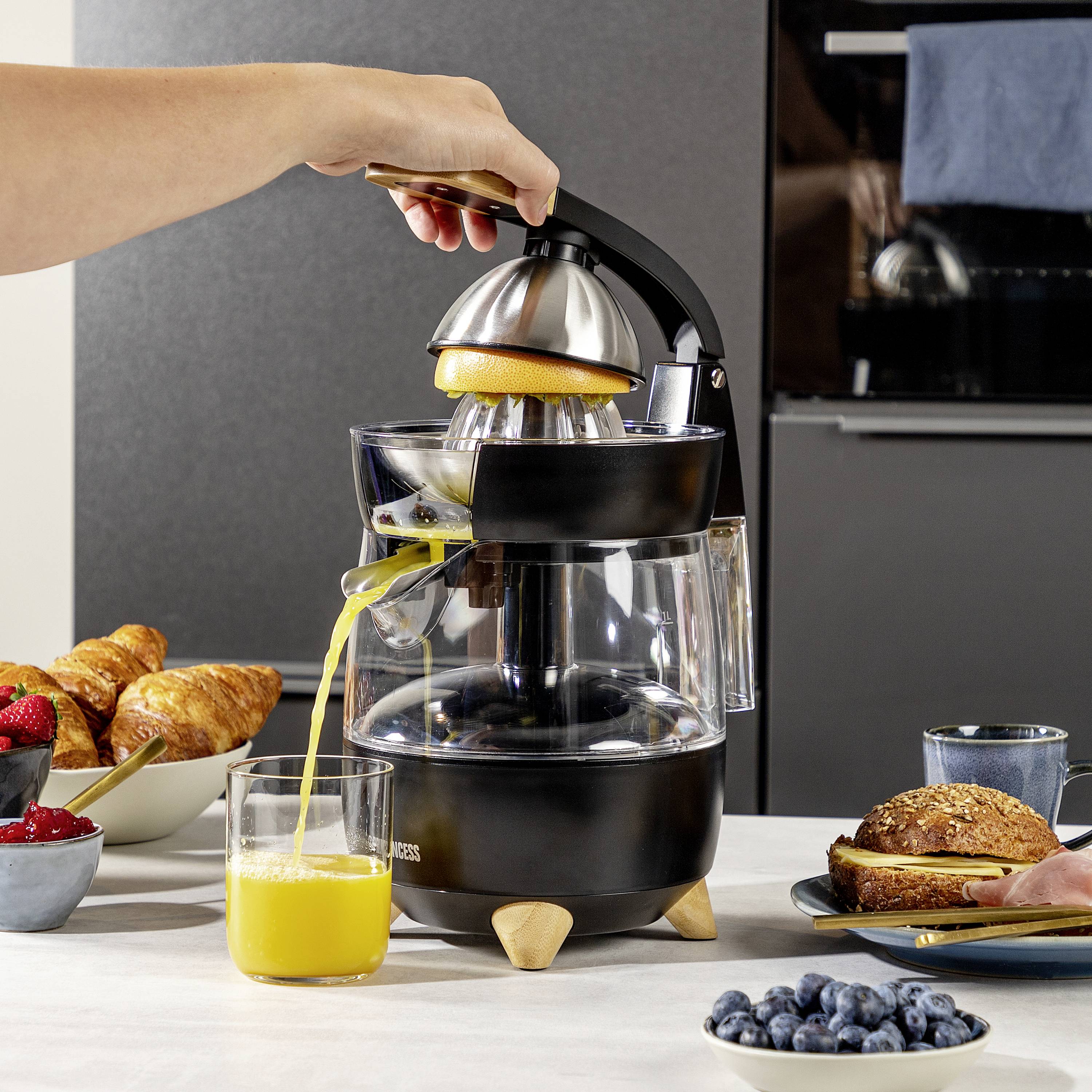 Eine Person presst eine Orange mit einer schwarzen Saftpresse aus. Vorne steht ein Glas mit frisch gepresstem Saft. Im Hintergrund Frühstück mit Brötchen und Beeren.