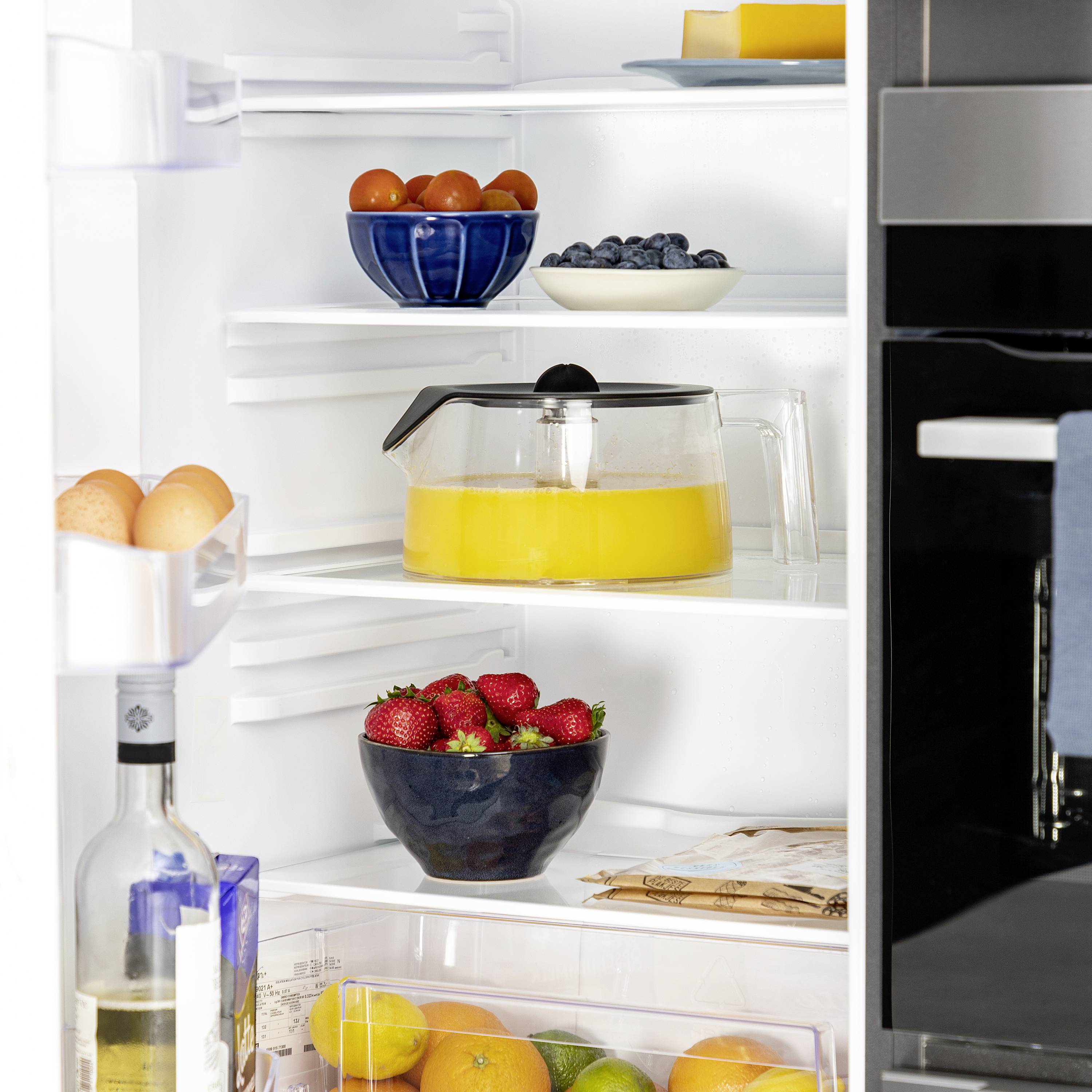 Ein Kühlschrank mit geöffneten Türen. Innen sind Orangen, Erdbeeren, Blaubeeren, Eier, eine Karaffe Orangensaft und ein großer Wasserkrug zu sehen.