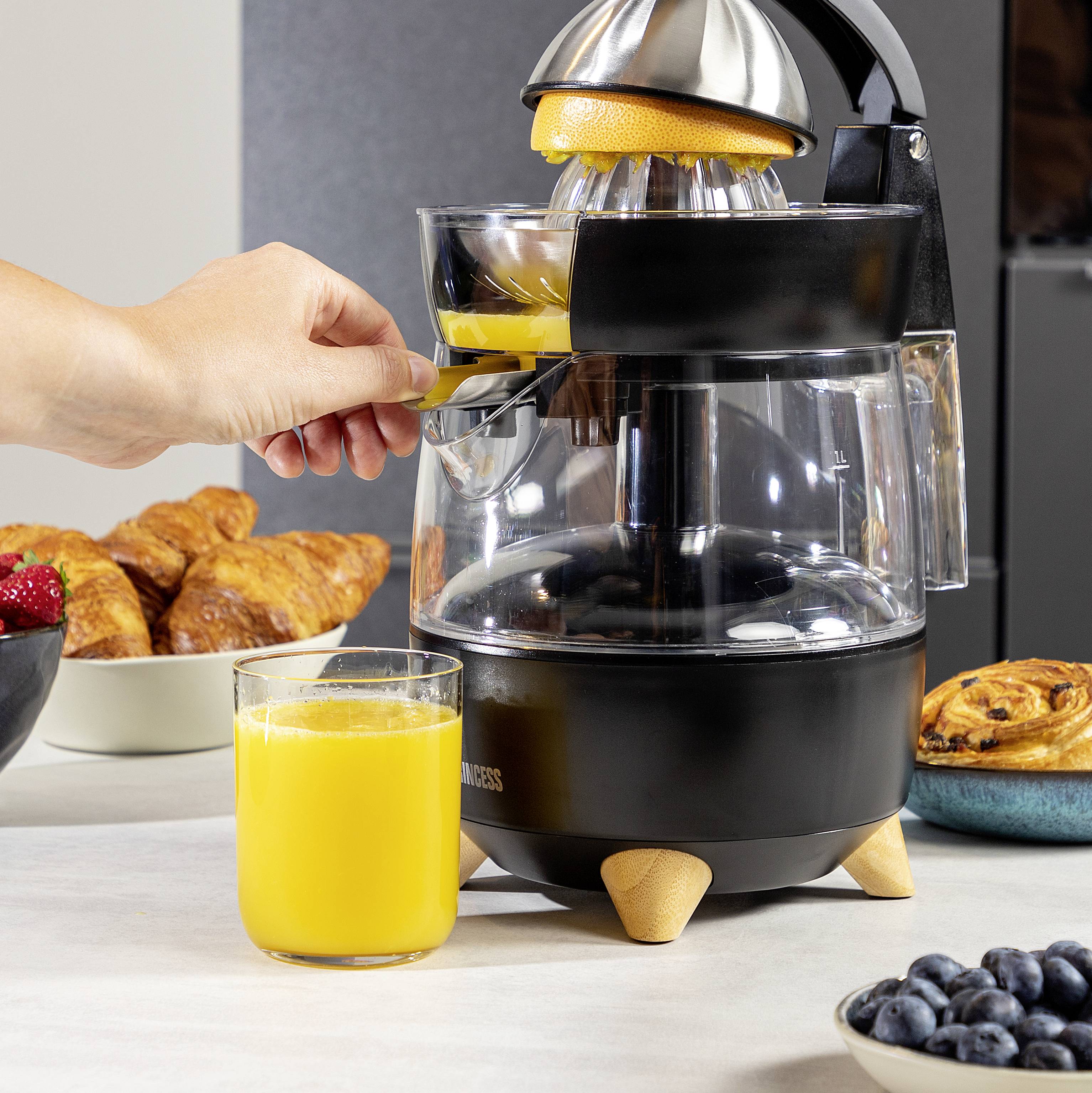 Eine Hand presst Saft aus einer Orange mit einer elektrischen Zitruspresse. Im Vordergrund steht ein Glas mit frisch gepresstem Orangensaft, im Hintergrund Croissants und Beeren.