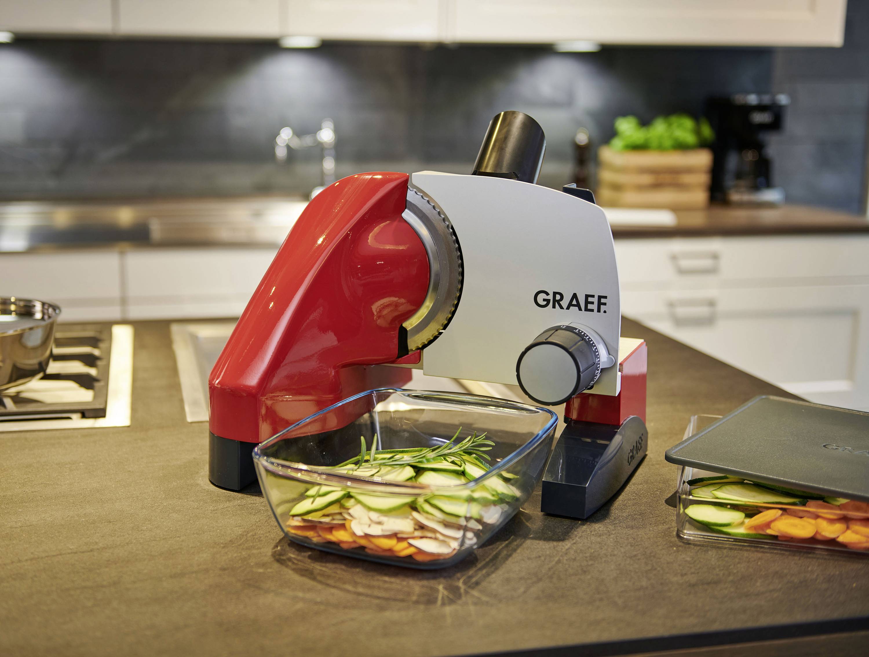 Eine rote und weiße elektrische Brotschneidemaschine steht auf einer Küchenzeile. Darunter befindet sich eine Glasschale mit geschnittenem Gemüse.