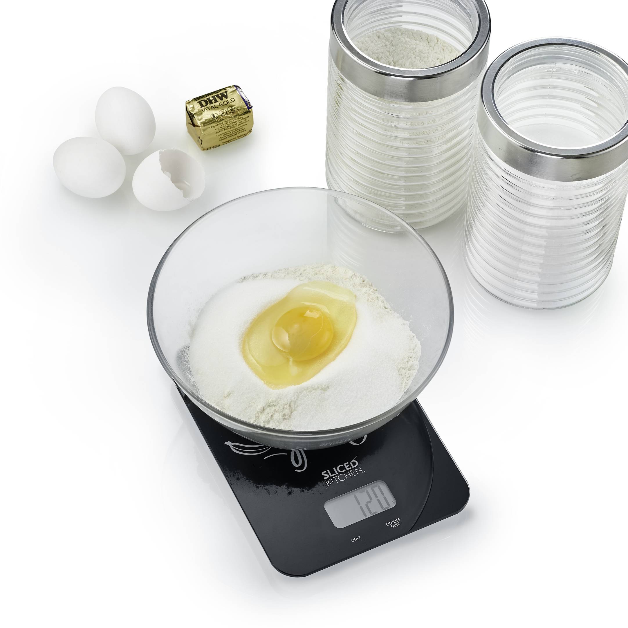 Eine Küchenwaage zeigt 128g Mehl mit einem Ei darauf. Daneben sind drei Eier, ein Stück Butter und zwei Behälter mit Zutaten.