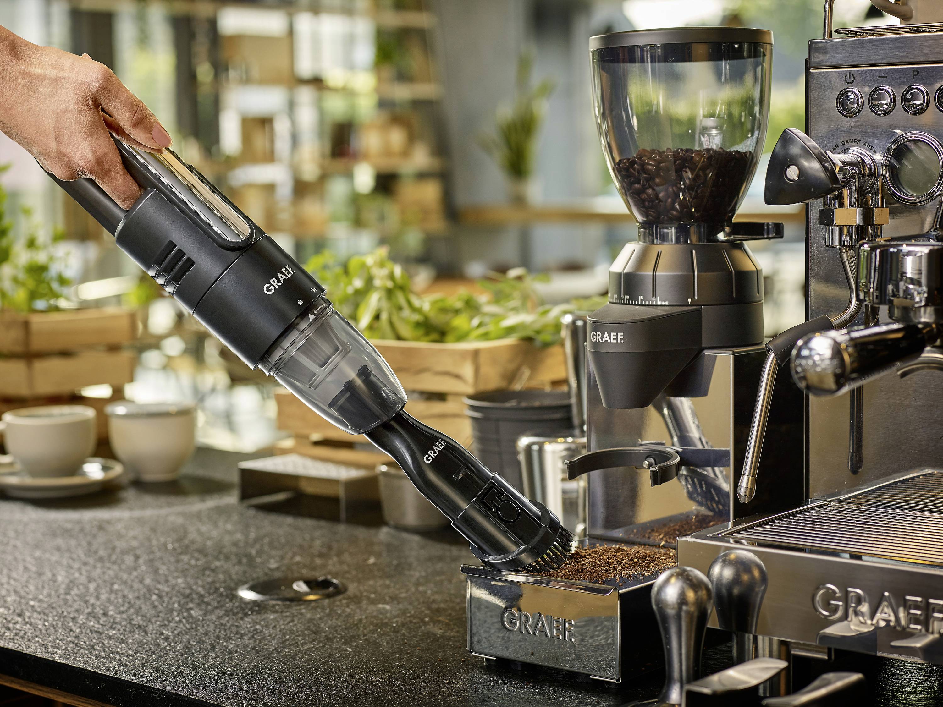 'Eine Handstaubsauger saugt Kaffeemehl neben einer Kaffeemaschine auf. Im Hintergrund sind Pflanzen und Geschirr zu sehen.'