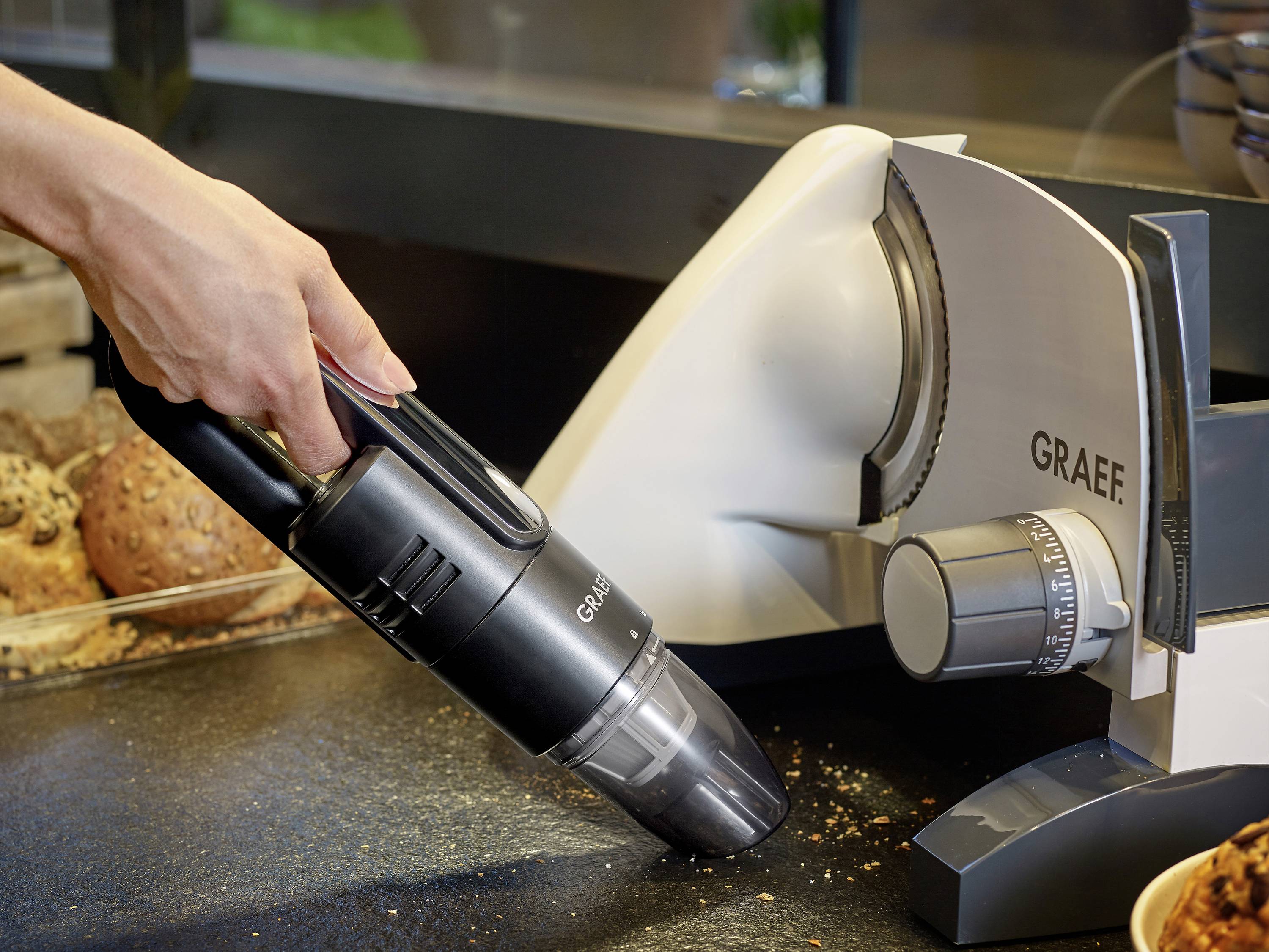 Eine Hand hält einen kleinen Staubsauger, der Krümel um eine Brotschneidemaschine aufsaugt. Im Hintergrund sind Brötchen zu sehen.