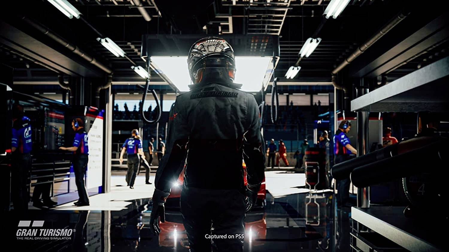 Ein Rennfahrer steht in einer beleuchteten Boxengasse, umgeben von Teammitgliedern und Fahrzeugen. Im Hintergrund ist Betriebsamkeit zu erkennen.