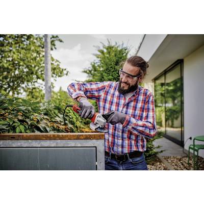 Ein Mann mit Bart und Schutzbrille schneidet mit einer elektrischen Heckenschere die Spitzen einer Hecke vor einem modernen Gebäude.