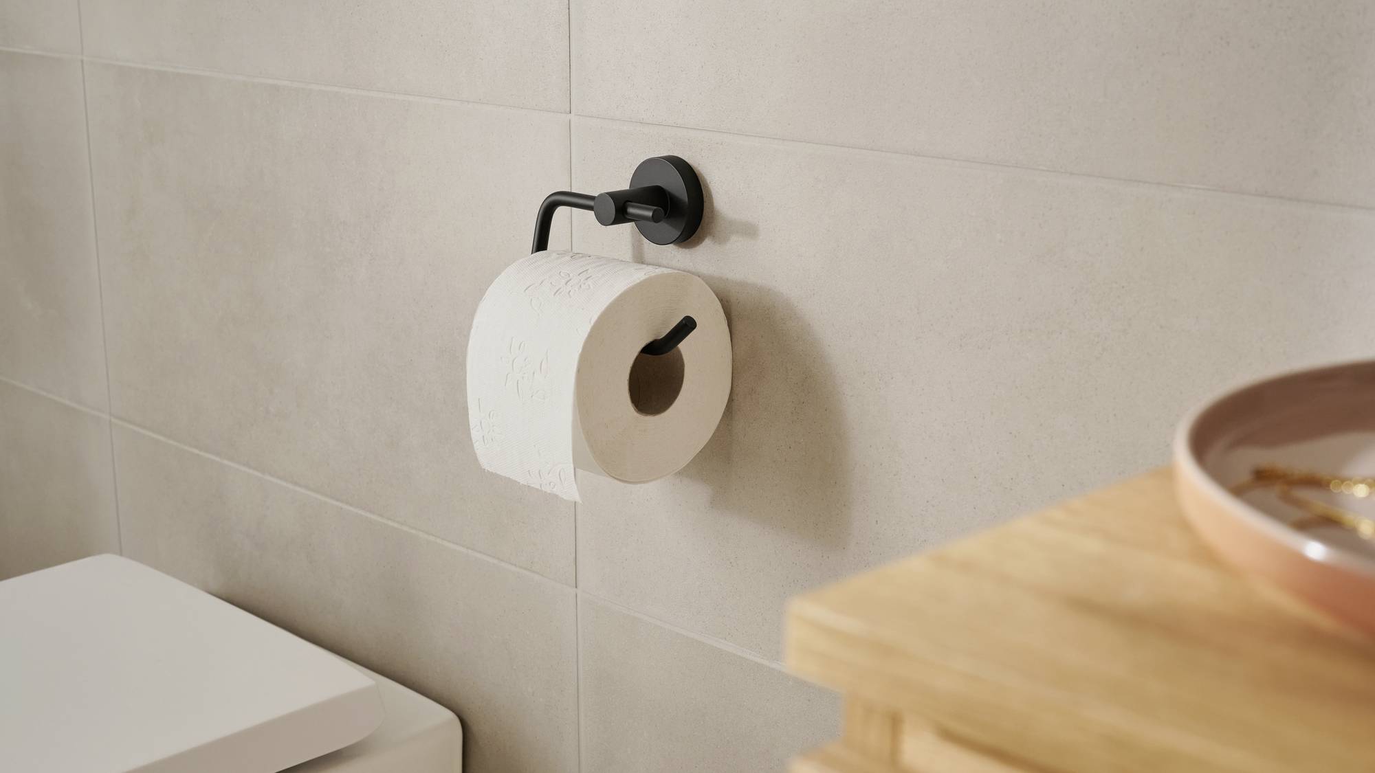 Toilettenpapierrolle hängt an einem schwarzen Halter an der Wand. Daneben befinden sich der Tank einer Toilette und eine Holzablage.