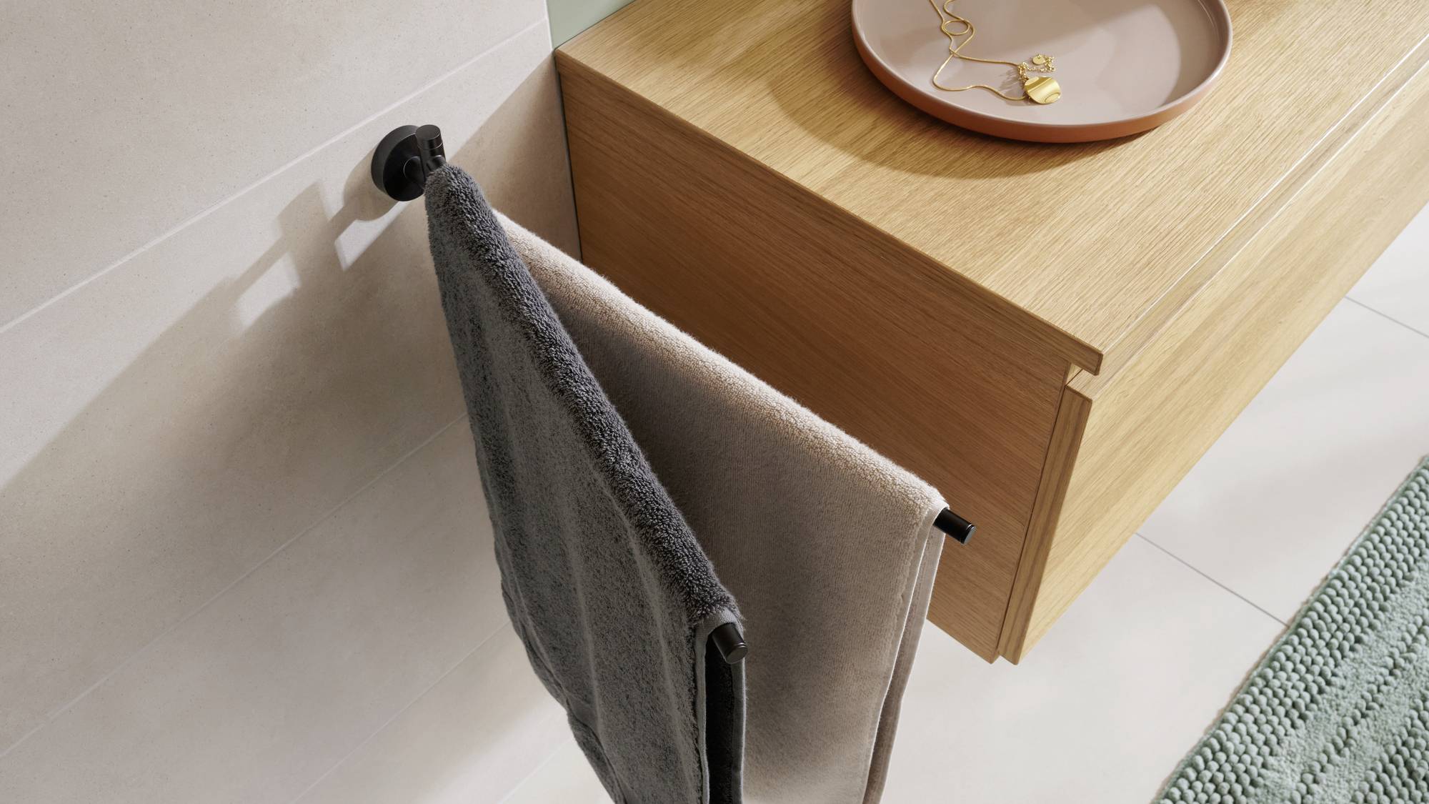 Handtuchhalter mit grauem Handtuch an einer Wand neben einer Holzmöbeloberfläche mit einem Teller und Schmuckstücken darauf.