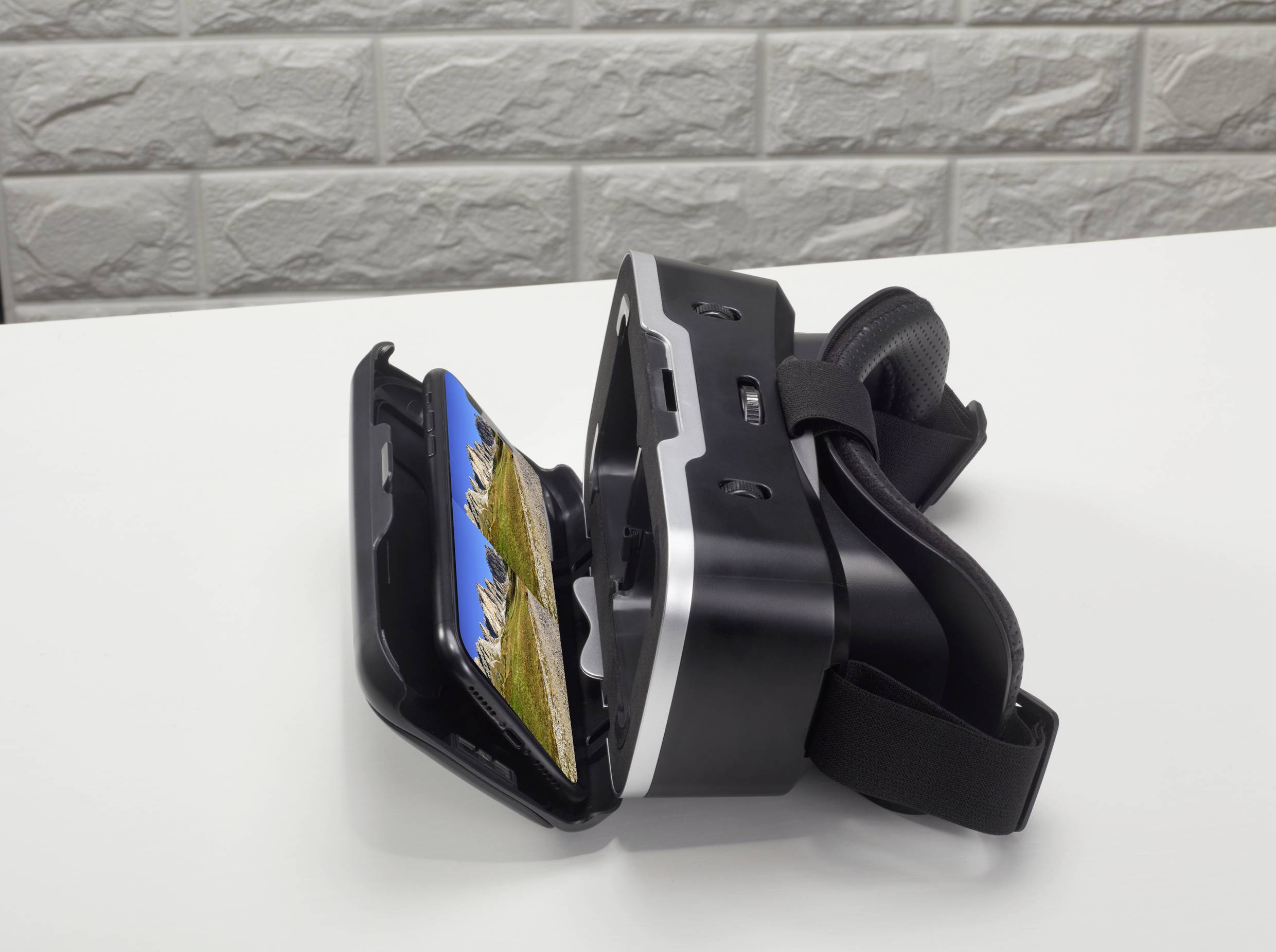 Virtual-Reality-Headset auf weißem Tisch, geöffnet mit einem Smartphone, das eine Berglandschaft anzeigt. Brickmauer im Hintergrund.